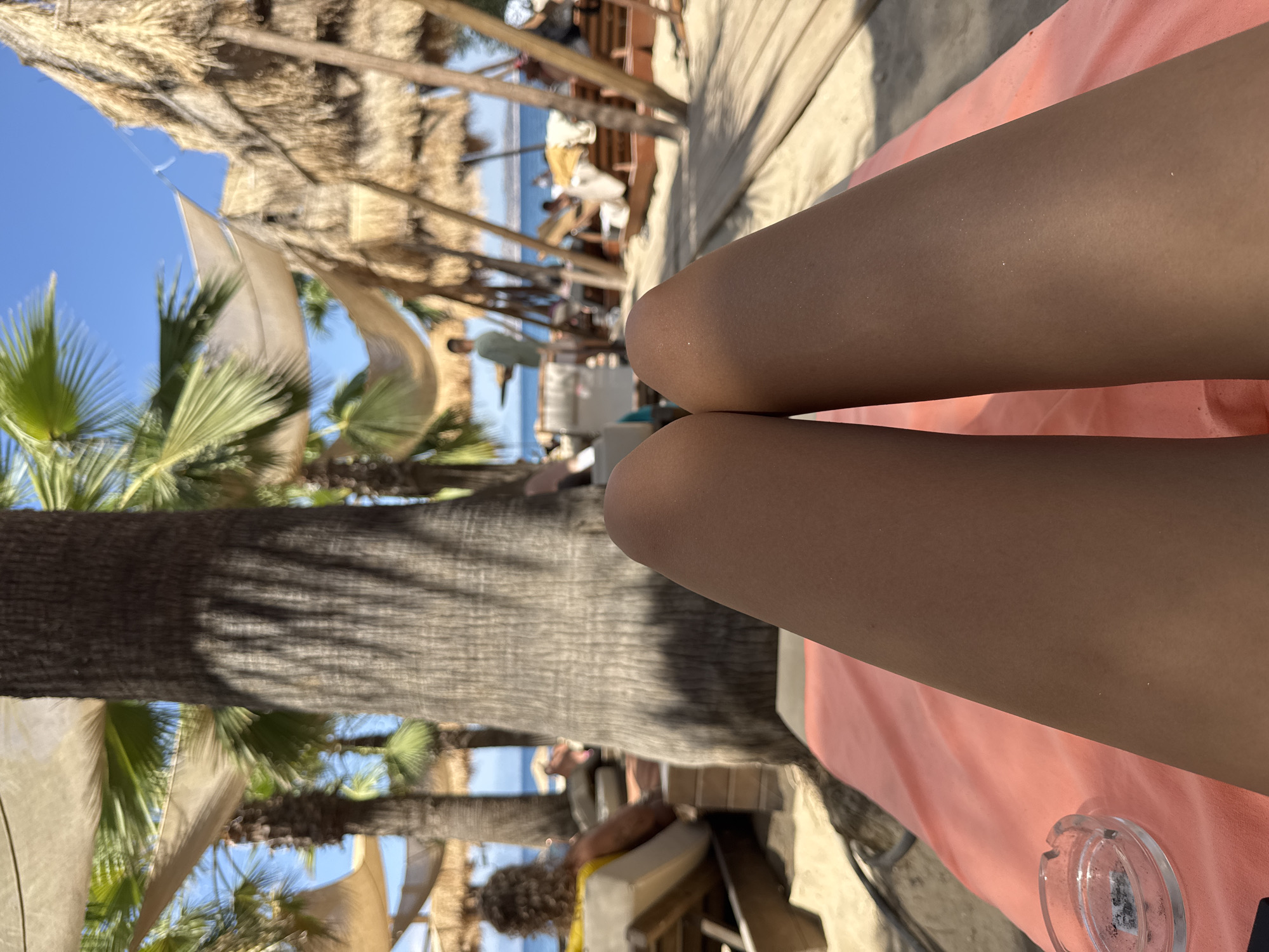 The image depicts a perspective from a beach chair on a sunny day in Thassos, Greece. The foreground is dominated by a person's tanned legs resting on a peach-colored towel. To the left, a glass ashtray with cigarette butts is visible. The background reveals a tropical beach setting with palm trees, thatched umbrellas providing shade, and lounge chairs occupied by other beachgoers. The blue sea and sky are visible in the distance. The overall scene conveys a relaxed atmosphere.
