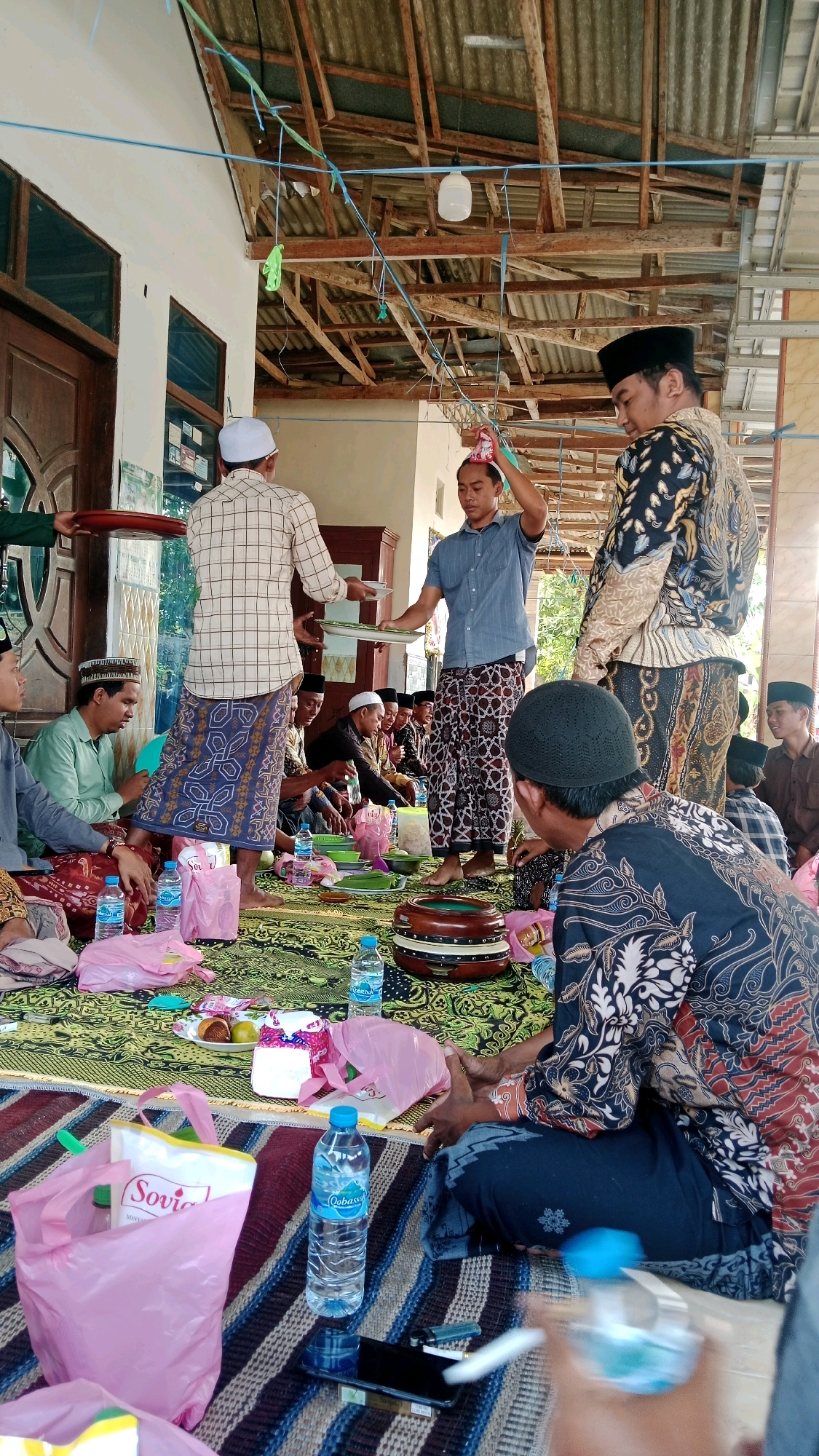 In Dempo Barat, Indonesia, a group of men are gathered on the floor, seated on a patterned mat and a woven rug. The setting appears to be an outdoor or semi-outdoor area with a visible wooden roof structure. Several men are dressed in traditional Islamic attire, including sarongs and skullcaps. Some are holding plates and food items, suggesting a communal meal or religious ceremony.
In the foreground, pink plastic bags and bottles of water are scattered on the ground. A large, circular musical instrument, possibly a rebana or kompang, is placed on the rug. Several individuals are engaged in conversation or observing the activities. The overall atmosphere seems to be one of social gathering and community.