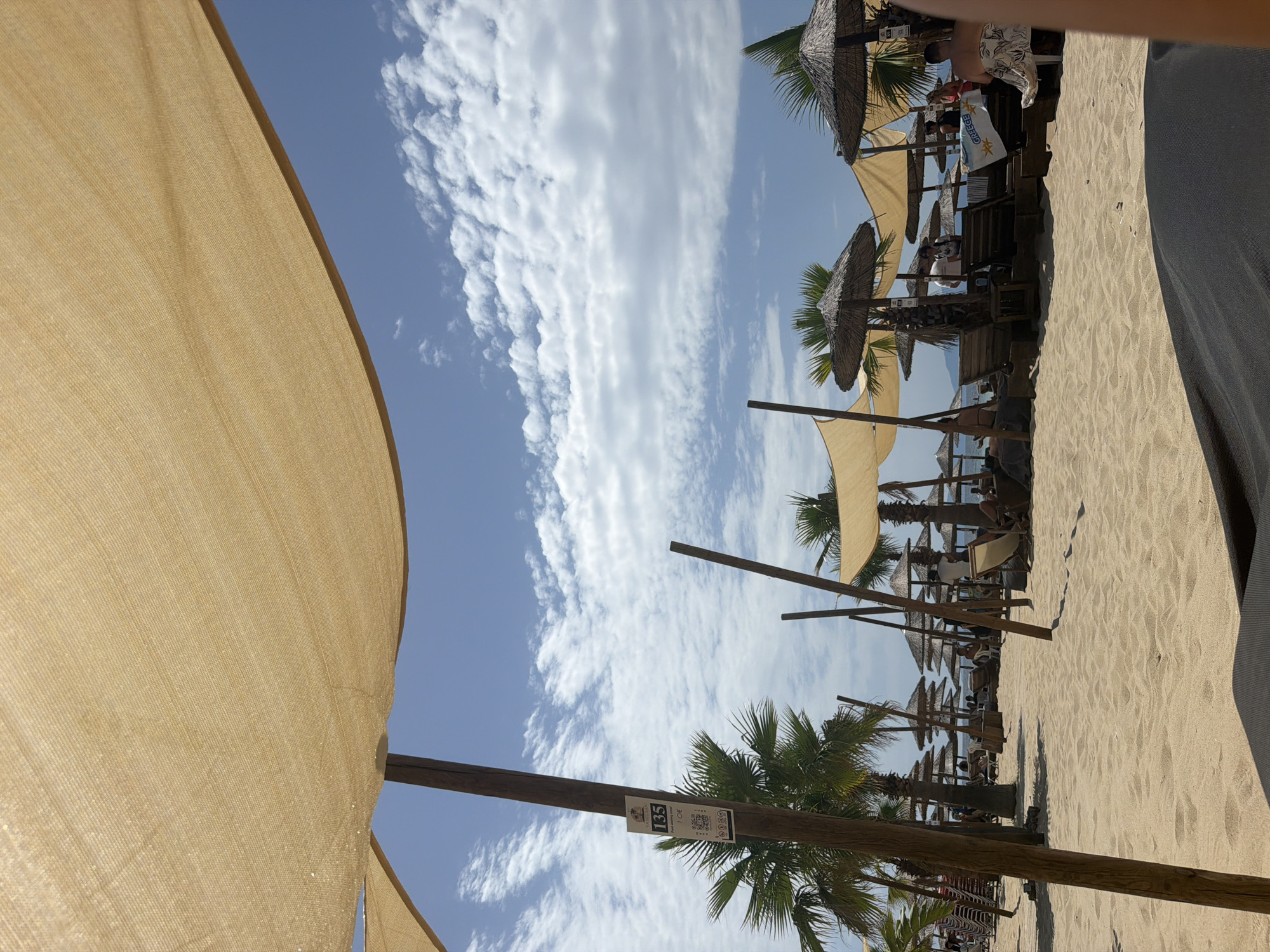 The image depicts a sandy beach with several thatched-roof umbrellas and lounge chairs set up. Palm trees are visible in the background, along with a clear blue sky with scattered white clouds. In the foreground, a large tan sunshade is draped over wooden poles, obscuring part of the view. A sign on one of the wooden poles reads "135" and "food waiting pool 1.0€". Several people are scattered around the beach, some sitting in chairs and others standing. A white flag with "GREECE" written on it is visible near the center of the frame. The overall scene suggests a relaxing atmosphere at a beach resort.