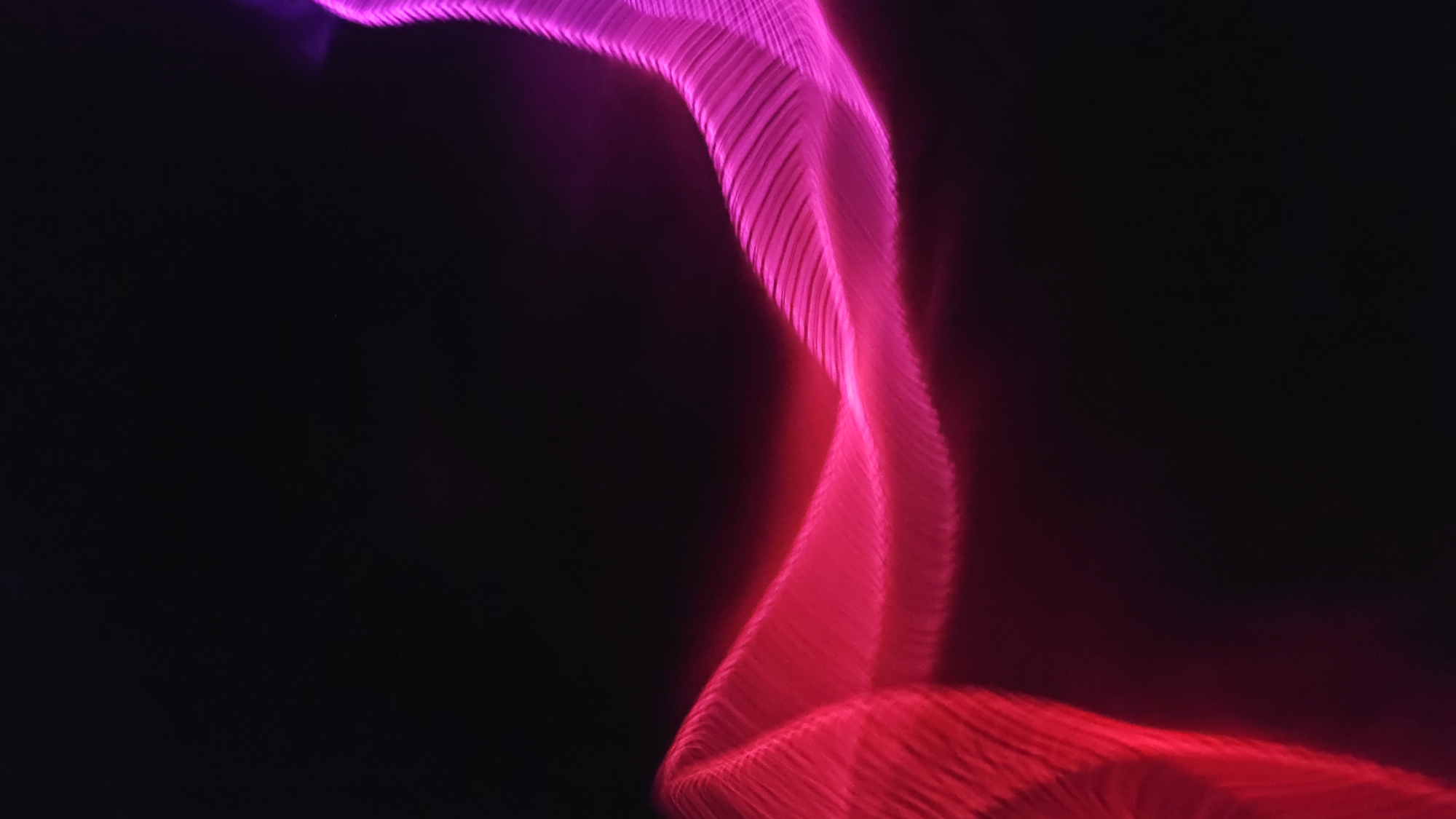 A long exposure photograph shows a twisting ribbon of light against a black background. The light ranges in color from deep red at the bottom to bright pink and violet at the top. The edges of the ribbon are blurred, and the light is striated, suggesting movement. The location in Cam Đức, Vietnam is not discernible from the image.