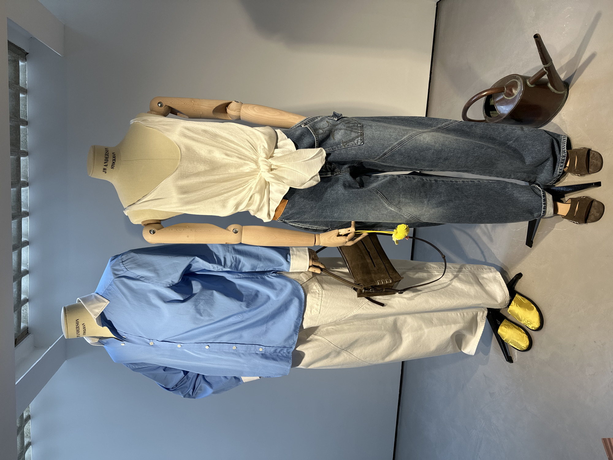 The image features two headless mannequins, displaying fashion ensembles in what appears to be a modern showroom or retail environment. The photograph itself is rotated 90 degrees clockwise, causing the upright mannequins and their surroundings to appear horizontally in the frame.
The mannequin on the right side of the image wears a cream-colored, pleated sleeveless top with a tied knot at the front, paired with dark wash, loose-fitting denim jeans. Its wooden, articulated arms are visible. It is accessorized with brown, open-toed flat sandals. Nearby, a vintage-style bronze watering can rests on the floor.
The mannequin on the left side of the image is dressed in a light blue long-sleeved button-down shirt with a white collar and cuffs, tucked into light beige or cream wide-leg trousers. Its wooden arms are articulated, and it holds a small dark brown shoulder bag from which a bright yellow flower emerges. The outfit is completed with bright yellow, open-toed flat mules.
The setting includes plain light grey walls and a matching floor, which has a smooth, concrete-like finish. A window with a grid pattern, possibly composed of glass blocks, is partially visible on the far left, allowing some light into the space. Both mannequins have "JW ANDERSON STOCKMAN" printed vertically on their neck area, indicating the brand or manufacturer. The overall scene depicts a static fashion display.