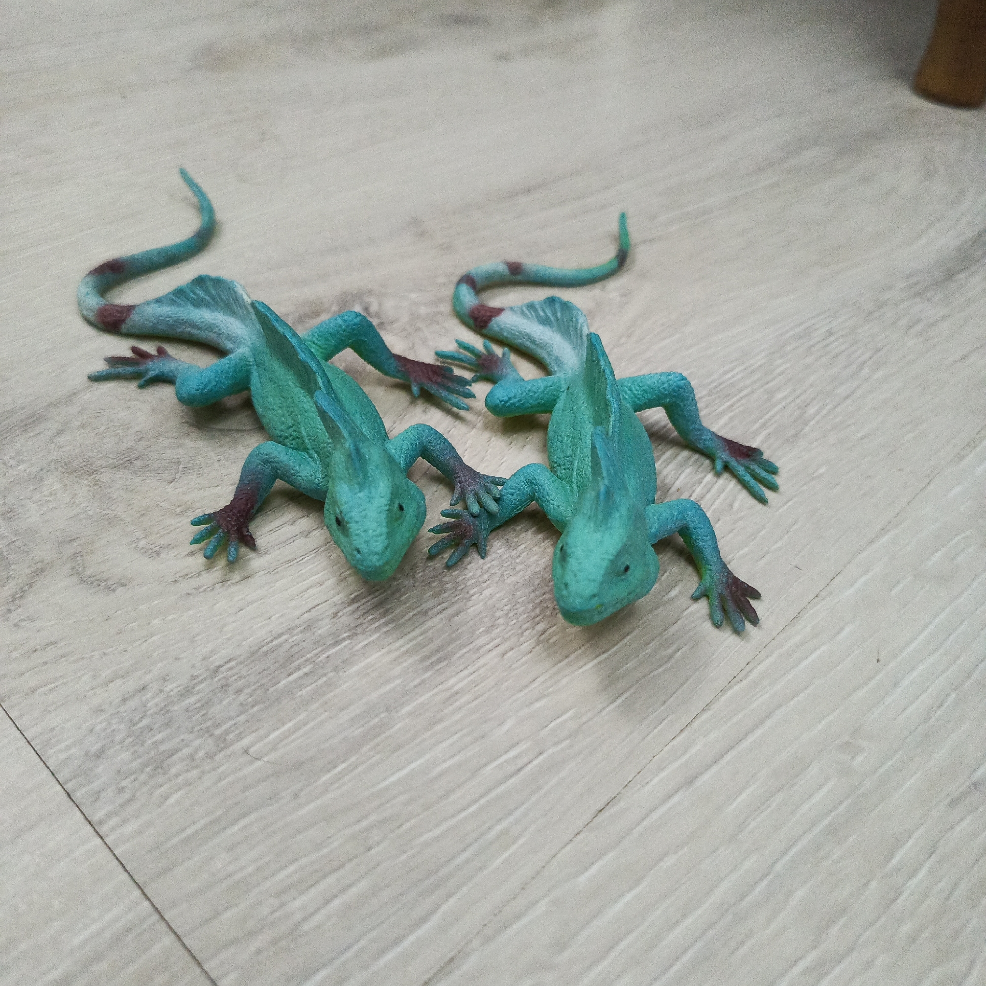 The image shows two turquoise toy lizards with brown markings on their tails, lying on a light-colored, wood-grain patterned surface. The lizards are positioned head-to-head, but slightly off-center. The environment is indistinct, suggesting a domestic setting, such as a child's playroom or living room. The image provides no information about the city of Марковский, Russia, or any people.