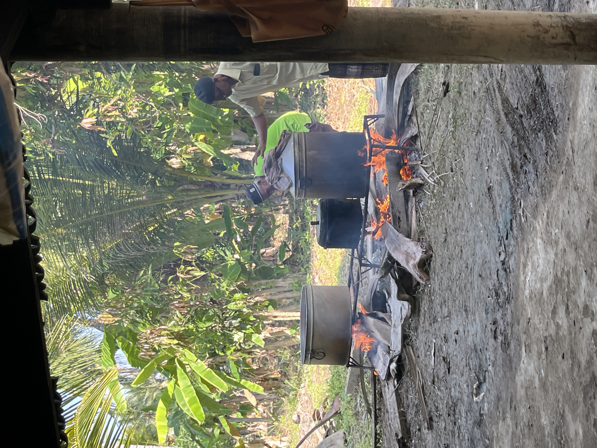 This image depicts an outdoor cooking scene in the village of Peurupok, Indonesia. Two men are tending to large metal pots set over open fires, which are fueled by wood and other debris. The area appears to be rustic, with a dirt ground surface and lush tropical vegetation, including banana trees and palm trees, in the background. One man, wearing a light-colored shirt and a black cap, is standing and interacting with the other man. The second man, dressed in a green shirt and a black cap, is leaning over one of the large pots, seemingly attending to the cooking process. A portion of a wooden structure with a dark roof is visible in the foreground, suggesting a covered area or shelter. The overall impression is one of traditional food preparation in a rural, tropical environment.