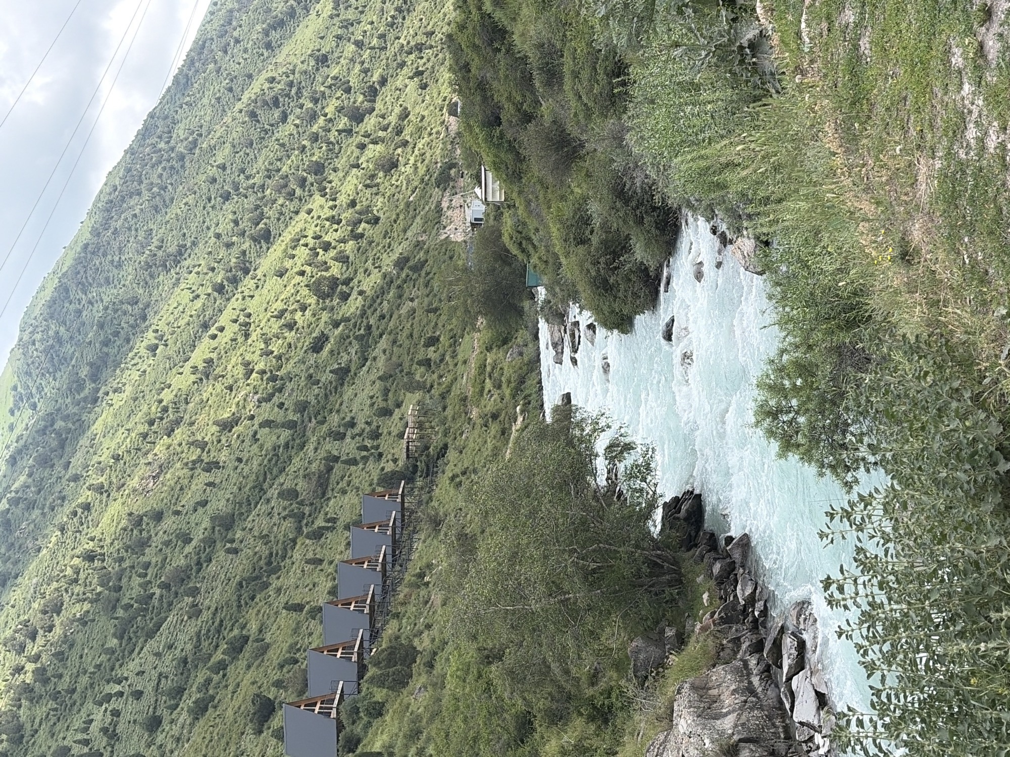 The image depicts a bright, daytime scene in a mountainous, natural environment. In the foreground, a turquoise river flows from right to left, with white rapids indicating swift movement. The riverbanks are lined with rocks and dense green vegetation. Across the river, a steep, green mountainside rises sharply, dotted with numerous evergreen trees. Along the upper part of the mountainside, a row of five triangular-shaped modern structures with dark roofs are visible, elevated on stilts. Further up the mountain, scattered buildings and power lines are present. The overall impression is one of a serene, somewhat remote location, possibly a resort or residential area integrated into a dramatic natural landscape.