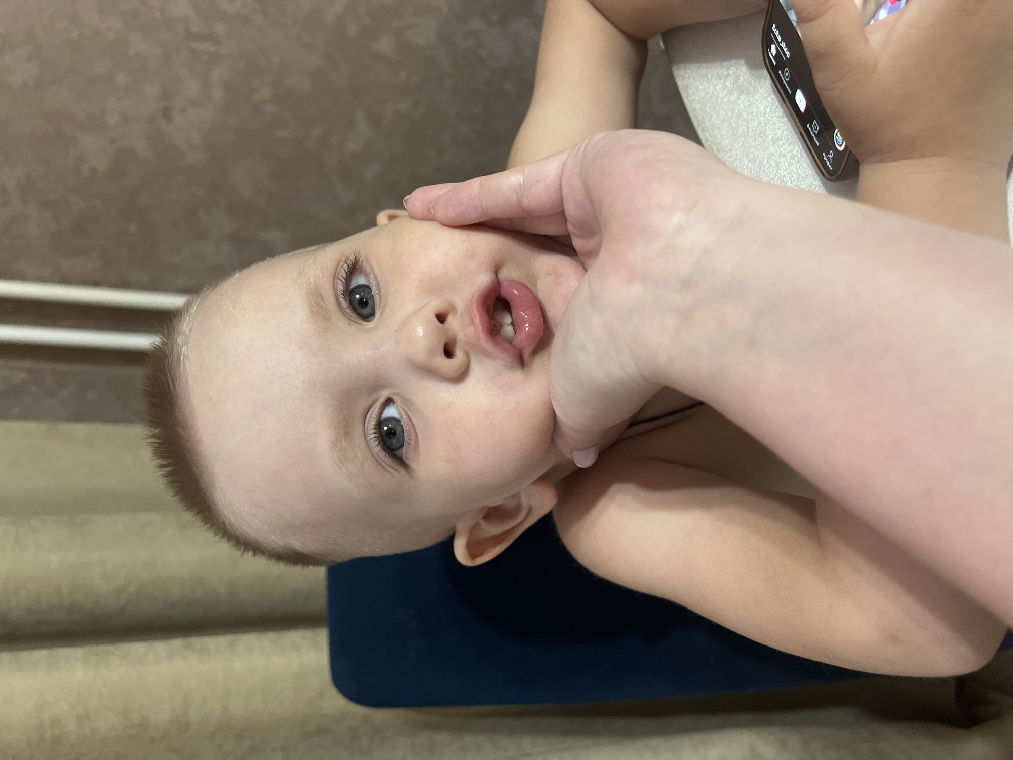A young, light-skinned child with short, reddish-blonde hair and blue eyes is partially visible in the image, their head resting on a blue surface. The child's mouth is slightly open, showing two front teeth. A light-skinned adult hand is touching the child's cheek, holding a smartphone. The background includes a textured, tan wall and a white radiator. The image was likely taken in the city of Марковский, Russia, but no objects or characteristics indicate this location.