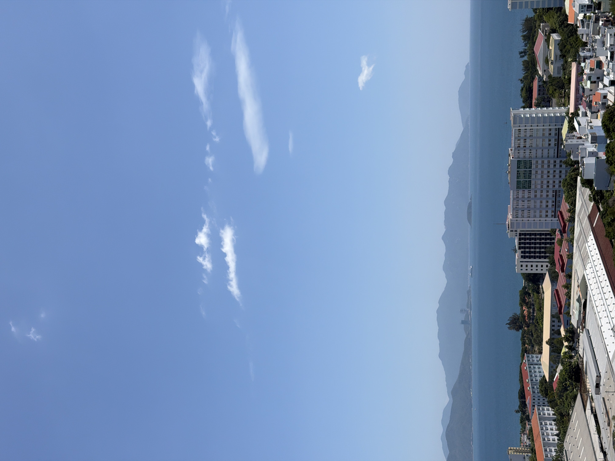 This is a vertical, eye-level, full shot from a high vantage point of the city of Nha Trang, Vietnam, on a clear day with a bright blue sky and sparse white clouds. The scene is dominated by the vast blue ocean in the mid-ground and a mountainous landscape in the far background.
In the foreground, the tops of several buildings are visible, some with red or orange tiled roofs and others with grey or white facades. These buildings appear to be part of an urban or industrial area. A large, light-colored, multi-story building with many windows and some green accents stands prominently in the mid-foreground. It is surrounded by lush green trees.
The ocean is a deep blue, stretching across the width of the image. Distant islands or peninsulas can be seen along the horizon, and on the far right, a white cruise ship is visible on the water. The mountains in the background are a hazy blue, suggesting they are quite distant.
The sky is a clear, vibrant blue, with a few scattered, wispy white clouds. No people are visible in the image. The overall impression is one of a coastal city on a sunny day, with a clear view of the sea and distant mountains.
