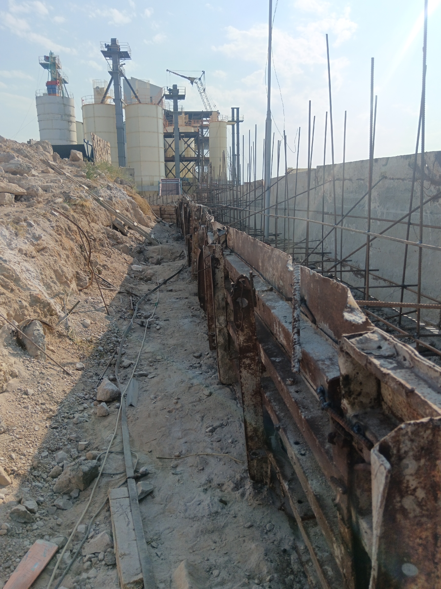 In Aragatsavan, Armenia, this construction site exhibits ongoing work on a foundation, likely for an industrial facility. In the background, several tall, cylindrical concrete silos and industrial structures suggest a cement plant or similar operation is nearby. A crane is visible, indicating heavy lifting and construction activities. The foreground shows a trench with rebar reinforcing structures and wooden or metal forms for concrete pouring. The ground is a mix of sand, rocks, and debris, with electrical wires and cables snaking along the trench. The scene is under a clear, bright sky, suggesting daytime. No people are visible in the frame.