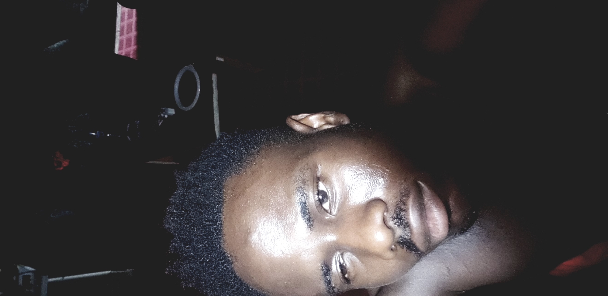 The image is a close-up, low-angle shot of a man's face, likely taken indoors in dim lighting. The man has dark skin, short black hair, a mustache, and appears to be lying down, possibly resting his head on a surface. His eyes are open. To the left of the man's head, in the background, are indistinct objects that appear to be part of a microphone setup, including a circular pop filter. There are also vague shapes indicating walls or other structures. The location in Narayi, Nigeria cannot be forensically confirmed from the image.