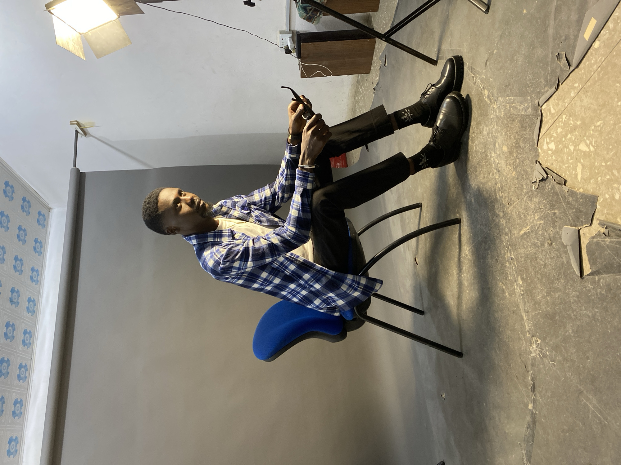 A man sits on a blue and black office chair in what appears to be a photography studio in Jos, Nigeria. He is wearing black pants, black shoes, black socks with white designs, and a blue and white plaid shirt. He is holding a dark-colored object, possibly a pipe or electronic device. The background is a gray wall with blue patterned wallpaper visible to the left. A light fixture is positioned above and to the left of the man. The floor appears to be concrete, with some damaged or peeling tiles visible. In the background, a cabinet is visible near a wall socket. Cables are visible throughout the scene.