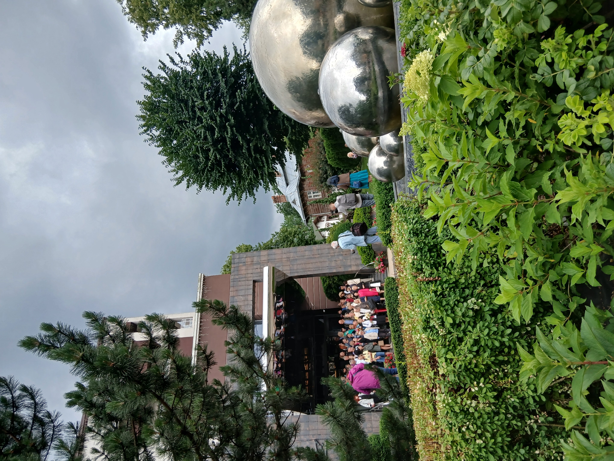 The image depicts a group of people gathered on the steps of a modern building with a dark, textured facade. In the foreground, lush green bushes and flowering plants are visible. To the right, a sculpture of several large, reflective spheres is present. The building appears to be the Grand Hotel Truskavets, situated in the city of Truskavets, Ukraine, a well-known spa resort. The sky is overcast with heavy clouds, suggesting a gloomy or perhaps rainy day. The group of people, composed of adults and possibly some children, are mostly looking towards the building or interacting with each other. Some are dressed in casual attire, while others appear to be in more formal wear. One person in the foreground, wearing a bright pink jacket, is facing away from the camera. The overall scene suggests an outdoor gathering or event taking place at the hotel.