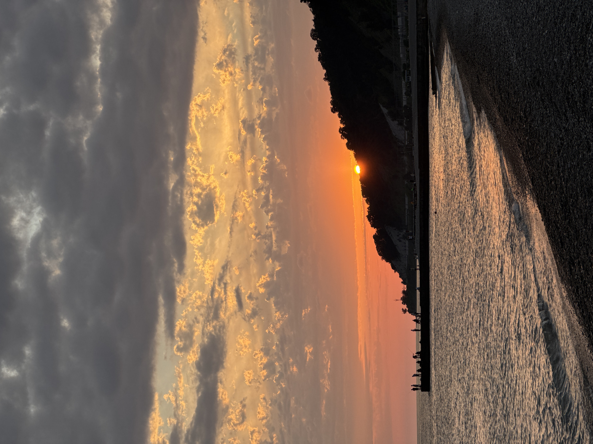 A sunset is captured over a body of water, possibly the sea, with a distinct shoreline. The sun is low on the horizon, partially obscured by a dark, tree-covered hill. The sky is filled with dramatic clouds, some dark gray and others illuminated with hues of orange and yellow from the setting sun. The water reflects the warm colors of the sky, with gentle waves lapping at the shore.
The shoreline itself appears to be made of dark pebbles. Along the edge of the water, a line of silhouetted figures can be seen, some standing and others seemingly fishing. These figures are small and indistinct, adding a sense of scale to the vastness of the scene. A dark, flat structure, perhaps a pier or breakwater, is visible running along the edge of the water, with the figures standing on or near it. In the distance, beyond the hill, there are hints of artificial structures, possibly buildings or lights, suggesting a populated area nearby.
The overall mood of the image is peaceful and serene, with the warm colors of the sunset creating a tranquil atmosphere. The time of day is clearly sunset, and the weather appears to be clear with scattered clouds. The location is a natural setting with a developed coastline. There is no visible text in the image.