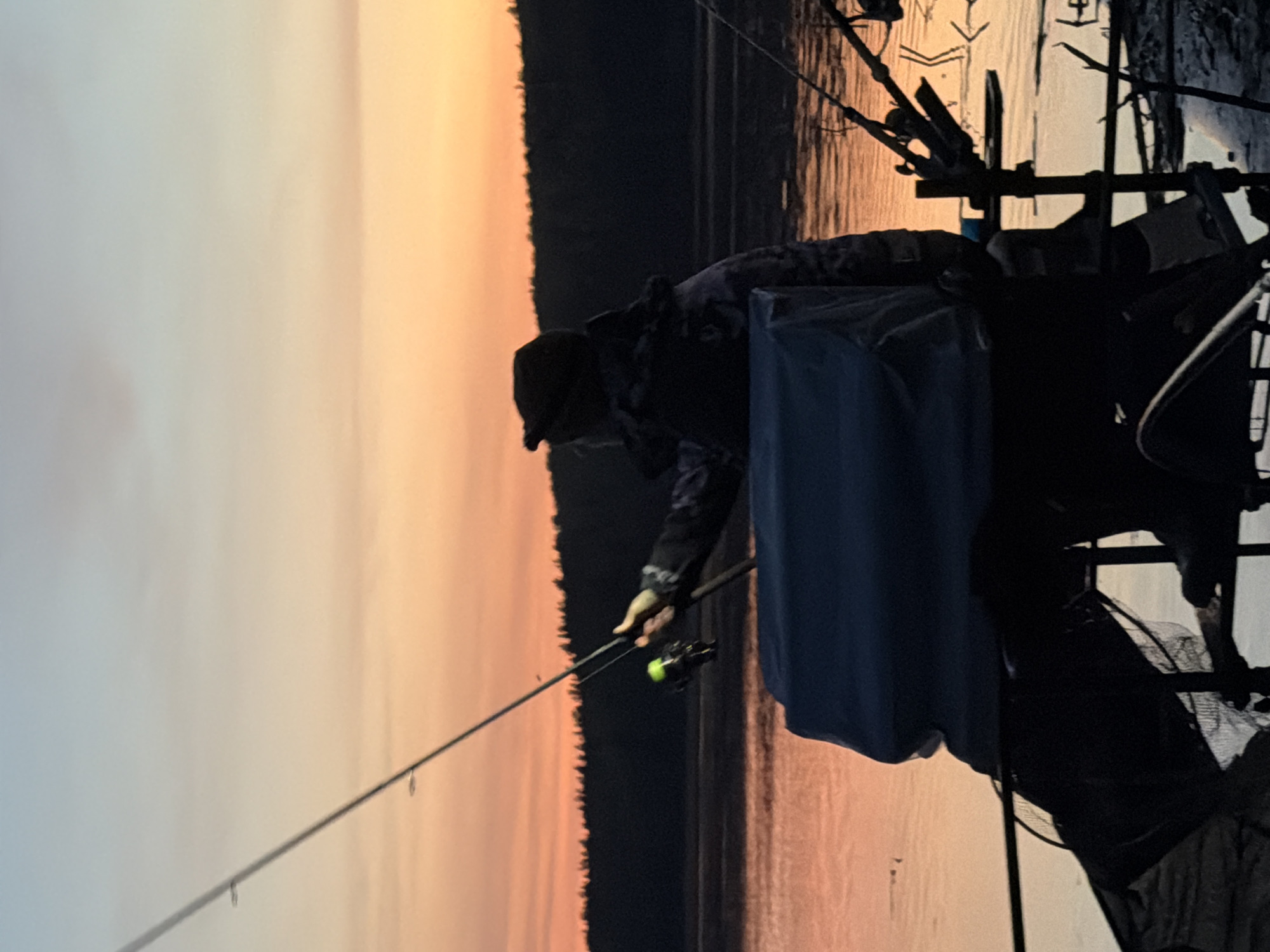 A person in dark clothing fishes at dusk. The sky is gradient, from pale at the left to dark orange near the edge of a dark forest. The person holds a fishing rod, the tip pointing out over the water. The person sits in a chair or platform with various accessories, rods, and containers. The water reflects the orange sky. The location and city are not evident in the image.