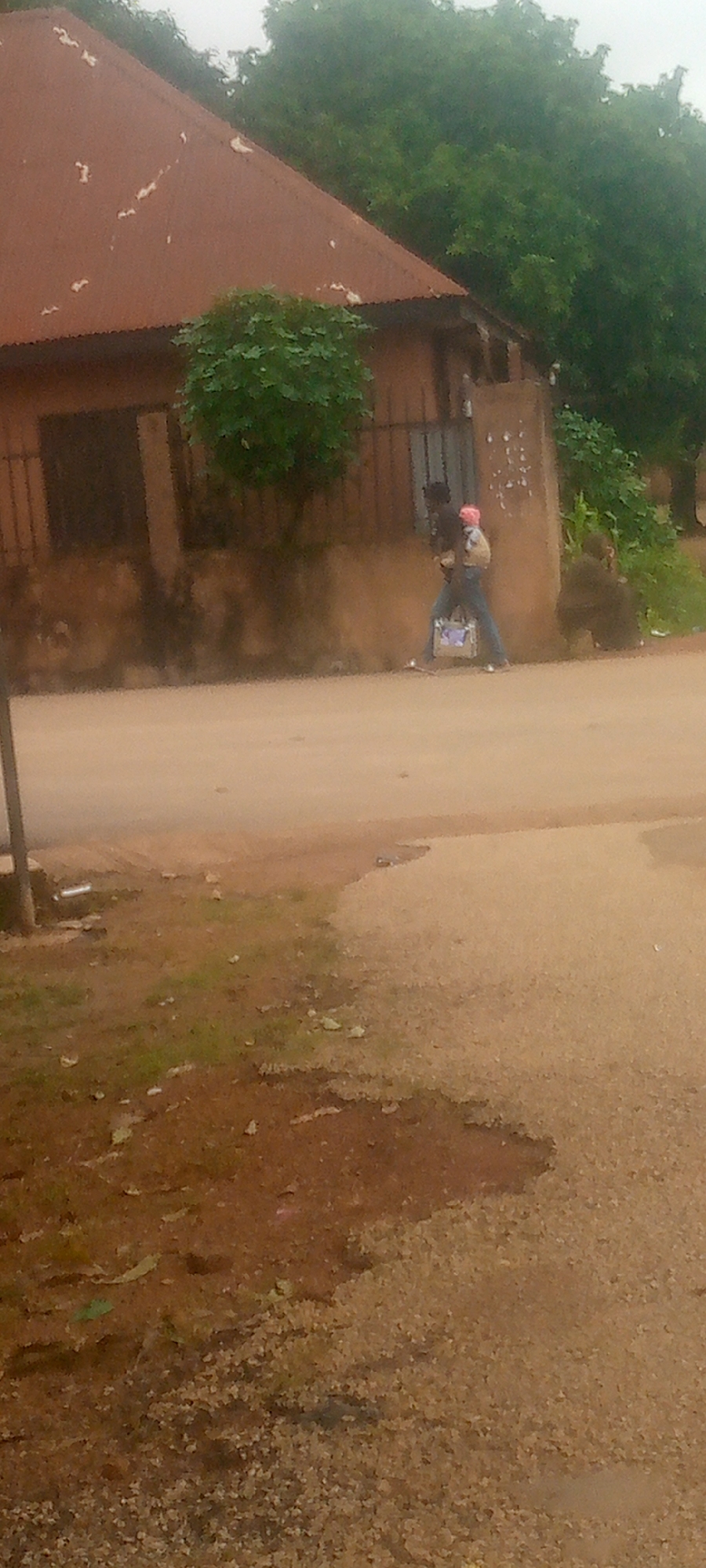 In an outdoor, dusty environment, two individuals are visible walking on a dirt road in front of a weathered, brown building with a rusted, corrugated metal roof. The building has barred windows and is partially covered by vegetation. The person in the foreground is carrying a light-colored bag. The person behind them appears to be carrying a child. The ground is covered in dirt and some sparse green vegetation. The sky is overcast.