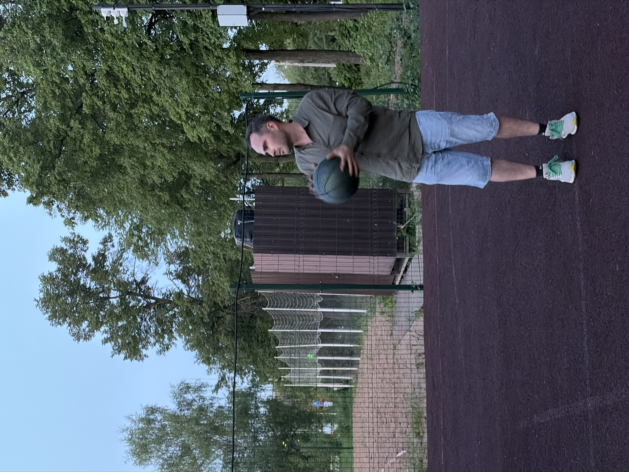 The image shows a man on a basketball court. He is wearing a dark green button-down shirt and light blue shorts. He is holding a basketball. Behind him is a fence, trees, and a building. The location is Izhevsk, Russia.
