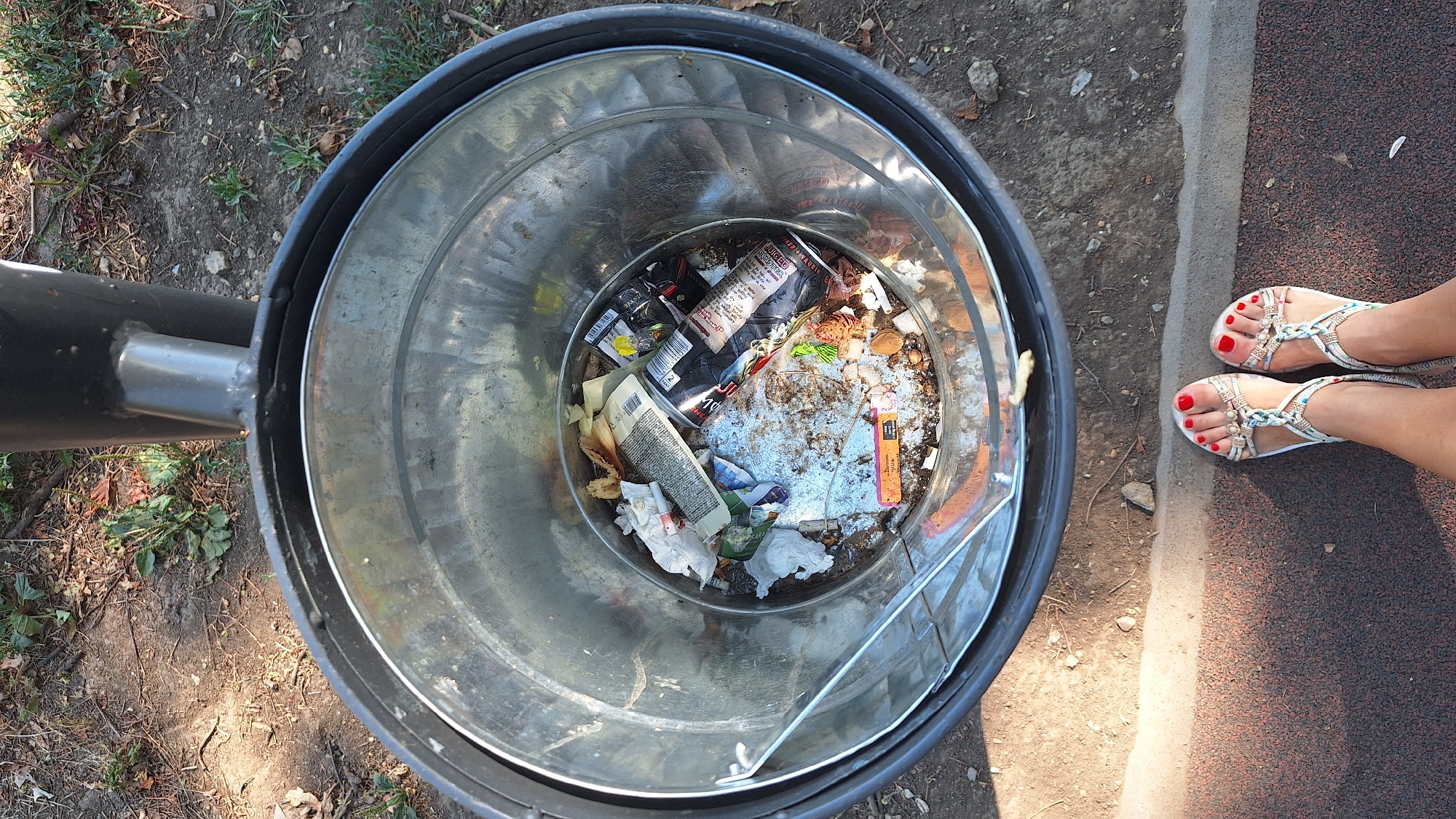 The image shows a close-up, overhead view of a metal trash can filled with various types of refuse. Among the visible items are crumpled paper, food wrappers, an empty aluminum can, and what appears to be cigarette butts. The trash can is positioned outdoors, with dry leaves and dirt visible on the ground surrounding it. In the foreground, a pair of feet wearing sandals is visible, suggesting a person is standing near the trash can. The overall scene appears to be an outdoor urban setting, possibly a park or public area. The location is identified as Sombor, Serbia.