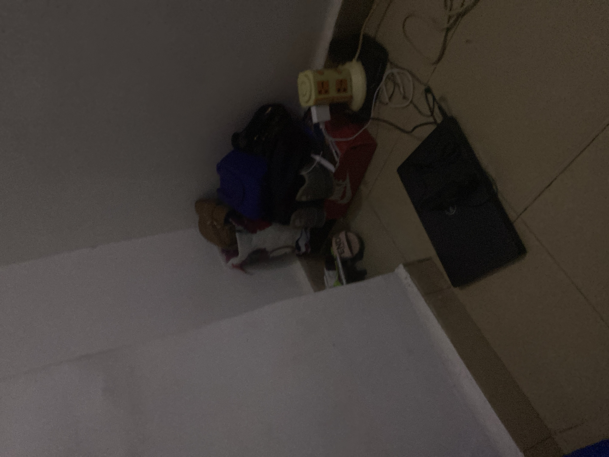 The image depicts a dimly lit corner of a room, possibly a residential space in Howayi, Nigeria. In the background, a collection of items is piled in the corner, including a blue duffel bag, a red box, a yellow cylindrical object, and various articles of clothing or fabric. A pair of sandals with the word "FEND" is visible on the floor near the pile. In the foreground, a black laptop is open on the tiled floor, connected to a power cord. The overall impression is one of a cluttered but lived-in space, with the focus on the utilitarian items and a hint of personal belongings.