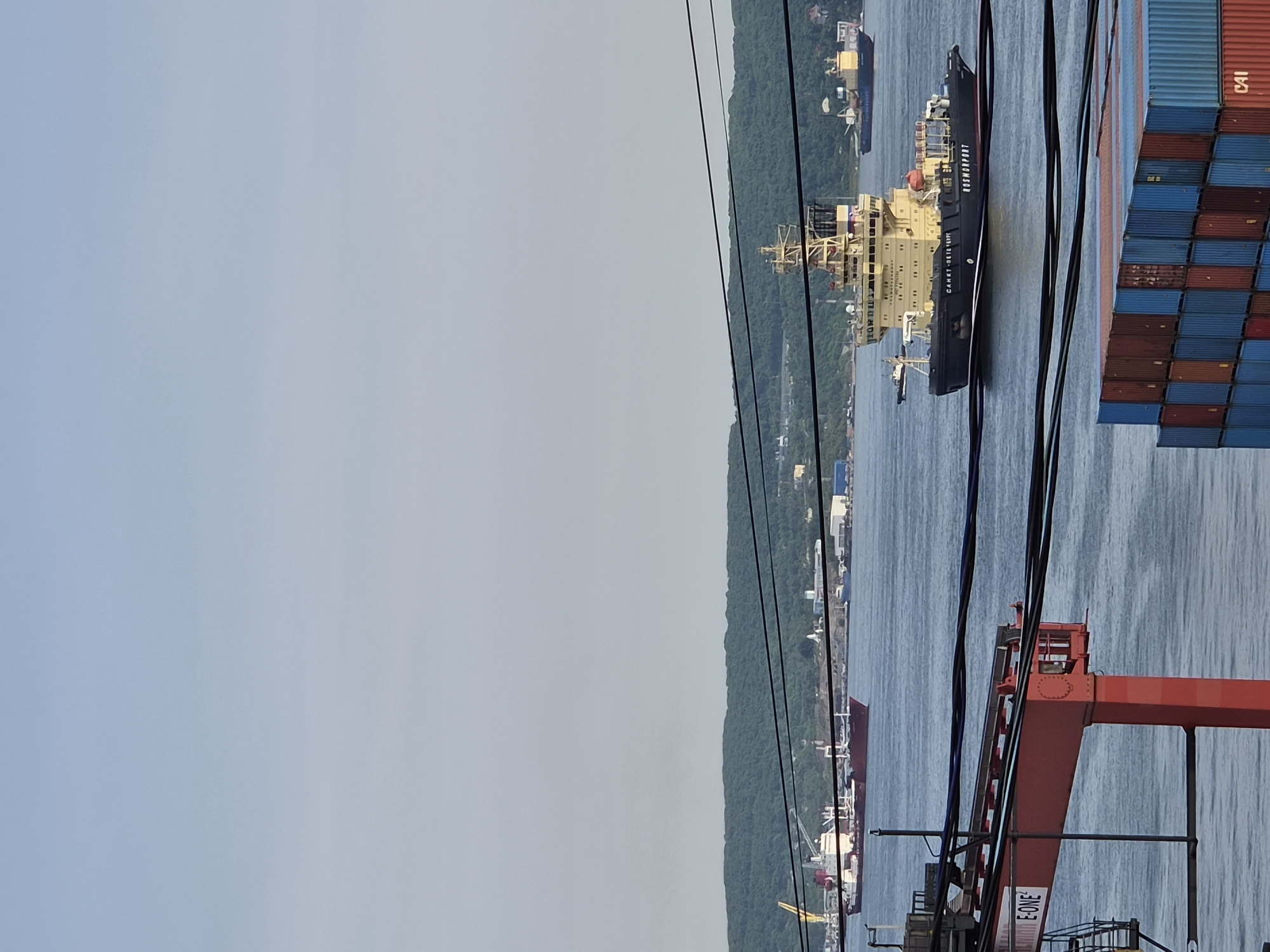 In Vladivostok, Russia, an icebreaker vessel named "KAPTAN DRANITSYN" with "ROSMORPORT" visible on its hull is navigating calm waters. The ship is dark with a yellow superstructure. In the background, a wooded hillside overlooks a coastal city with industrial buildings and other ships. In the foreground, a red port crane with "E-ONE" written on it is visible, along with stacks of shipping containers. The sky is clear and pale blue.