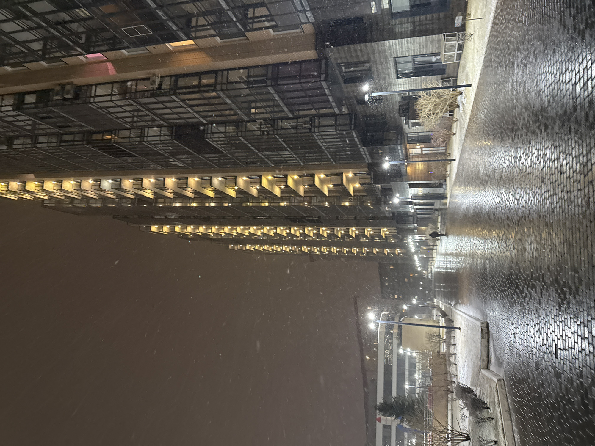 The image depicts a street scene in what appears to be a modern urban area, possibly in Krasnoyarsk, Russia. It is nighttime, and snow is falling. A long, multi-story building with rows of illuminated balconies runs along the left side of the frame. The street is paved with brick or cobblestone, and its surface is wet and reflective, likely from the snow. Streetlights are visible along the street. A solitary pedestrian is seen in the distance, walking along the sidewalk. There is another building with modern architecture visible further down the street. Sparse trees or bushes are also present along the street. The scene is generally bleak, with low visibility due to the snow and darkness.