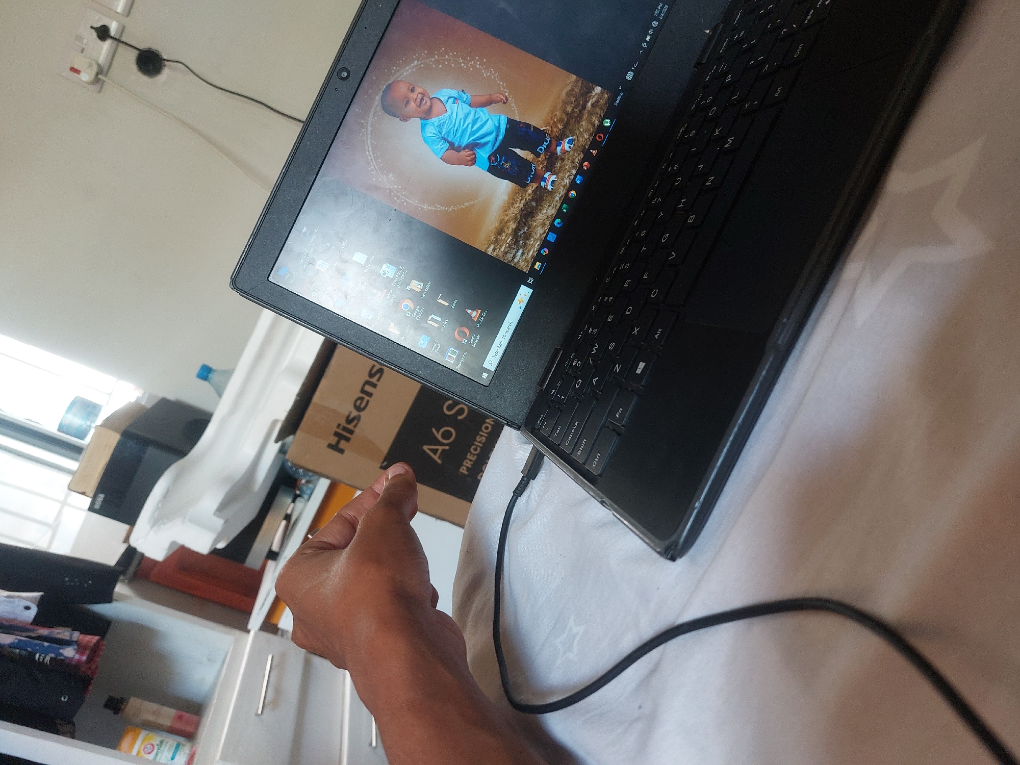 The image shows a black laptop resting on a light-colored surface, possibly a bed, with a child's image displayed on the screen. The laptop is connected to a charging cable. A hand is partially visible near the laptop, and a "Hisense A6 S PRECISION" box is partially in the background. Other objects like a drawer, a cabinet and bottles are also in the background. The location, Dar es-Salaam, Tanzania, is not evident from the image.