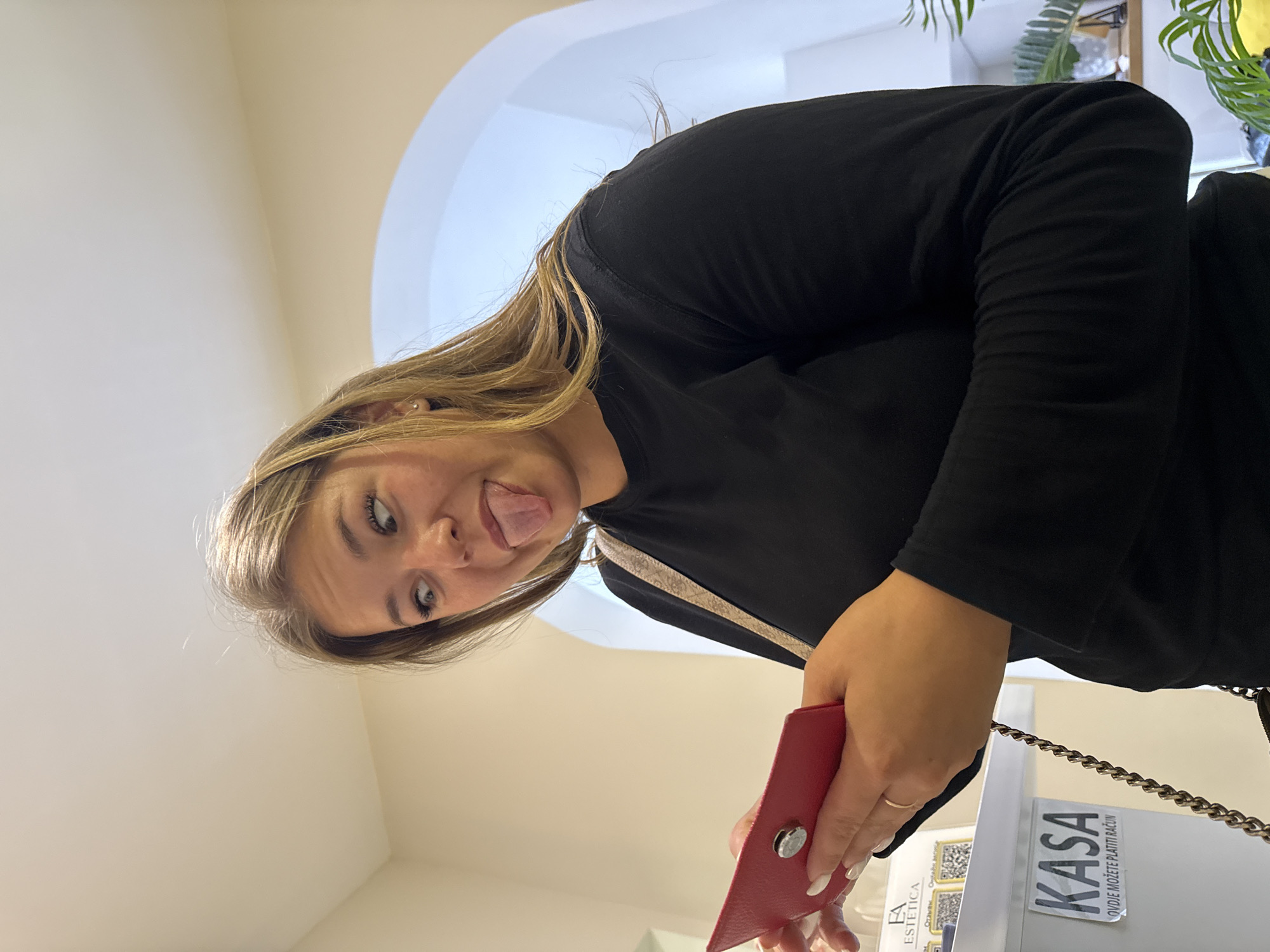 A woman with long, blonde hair is sticking her tongue out, holding a red wallet, and standing near a "KASA" sign. The sign reads "OVDJE MOŽETE PLATITI RAČUN". The location is likely in Budva, Montenegro.