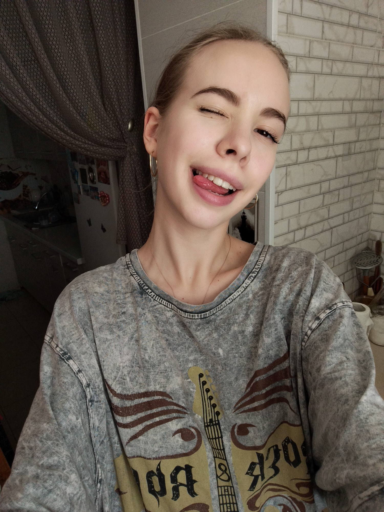 A young woman with blonde hair pulled back in a bun is taking a selfie. She is winking and sticking her tongue out, with her mouth open and teeth showing. She is wearing a gray, acid-wash t-shirt with a graphic print on the front that appears to be a guitar with some lettering. She has a thin gold necklace on. The background is blurry, but shows a kitchen or dining area with curtains on the left and a tiled wall on the right. There is a refrigerator visible in the background.
