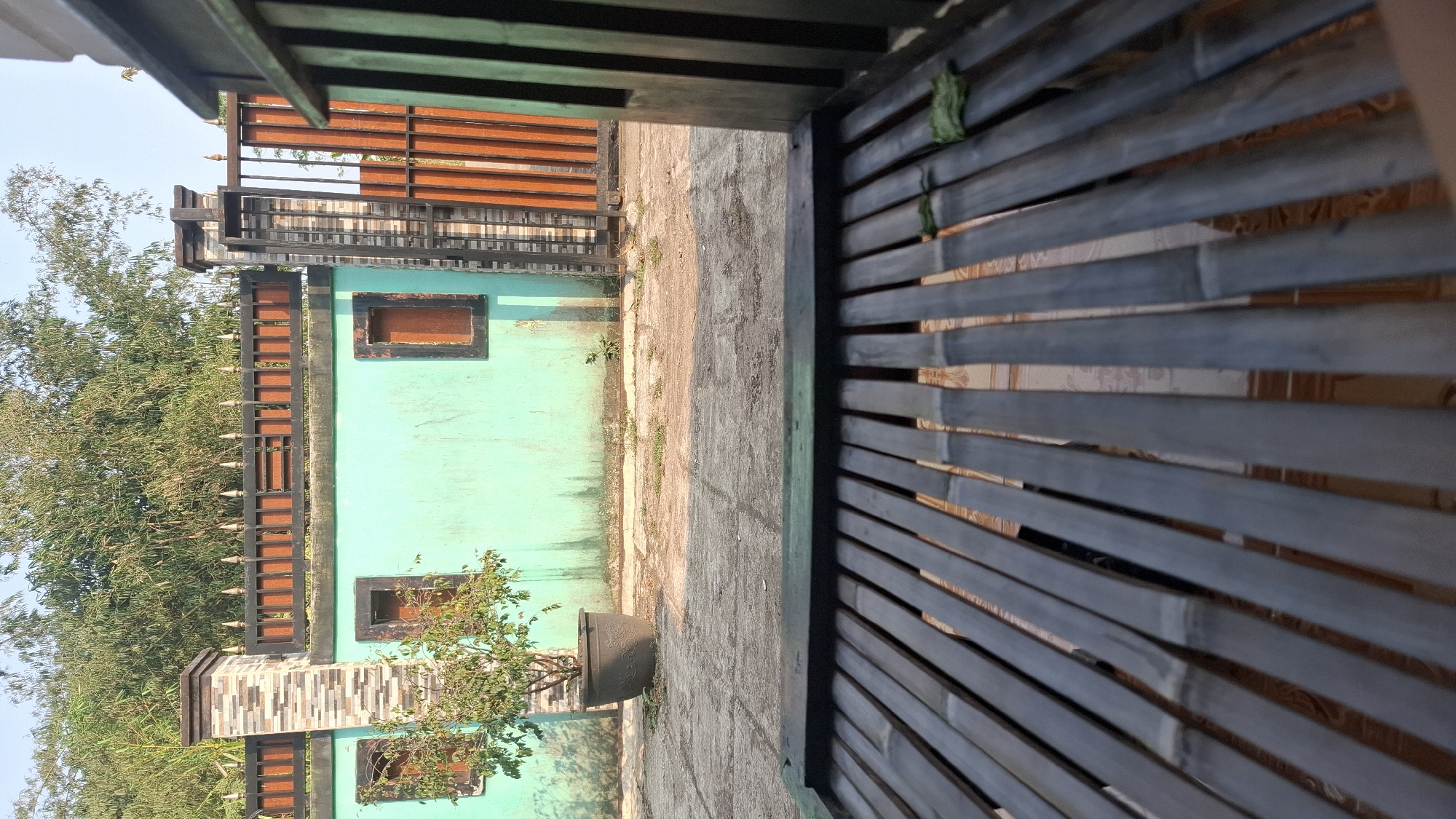 The image shows a tranquil outdoor scene in Cibinong Hilir, Indonesia. In the foreground, a dark-colored bamboo bench with a woven surface is positioned, suggesting a relaxed seating area. Beyond the bench, a light blue wall with three small, square windows, each framed with dark wood and containing a reddish-brown interior, stretches across the midground. To the left of the wall, a single potted plant with green foliage adds a touch of nature. A decorative chimney or pillar made of stacked rectangular tiles, transitioning from grey to brown and beige, stands near the left side of the wall.
The wall is topped with a black metal fence featuring a repeating pattern of vertical bars and horizontal supports, with pointed finials along the top edge. To the right, a gate made of vertical orange-brown slats is visible, also part of the fence. Behind the wall and fence, lush green bamboo foliage creates a natural backdrop. The ground in front of the wall appears to be a paved area with visible cracks and some sparse vegetation growing through. The scene is illuminated by natural sunlight, casting shadows on the ground, indicating it is daytime. The overall atmosphere is one of calm and simplicity, characteristic of a residential outdoor space.