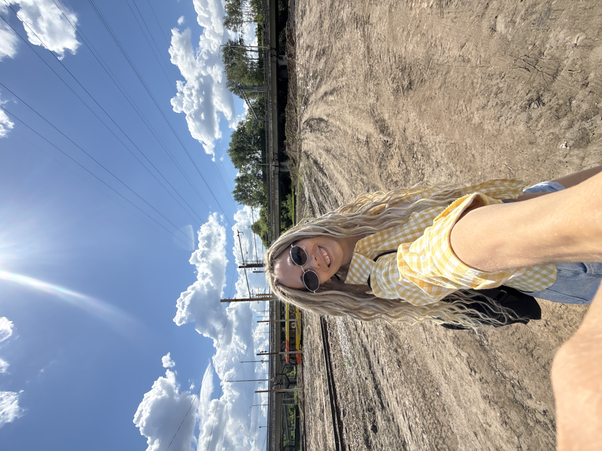 A selfie shows a woman wearing sunglasses and a yellow gingham shirt, standing next to train tracks under a blue sky with puffy white clouds. Power lines and a bridge structure are visible in the background. The ground is dirt and gravel. The picture was taken outdoors in Заволжье, Russia.