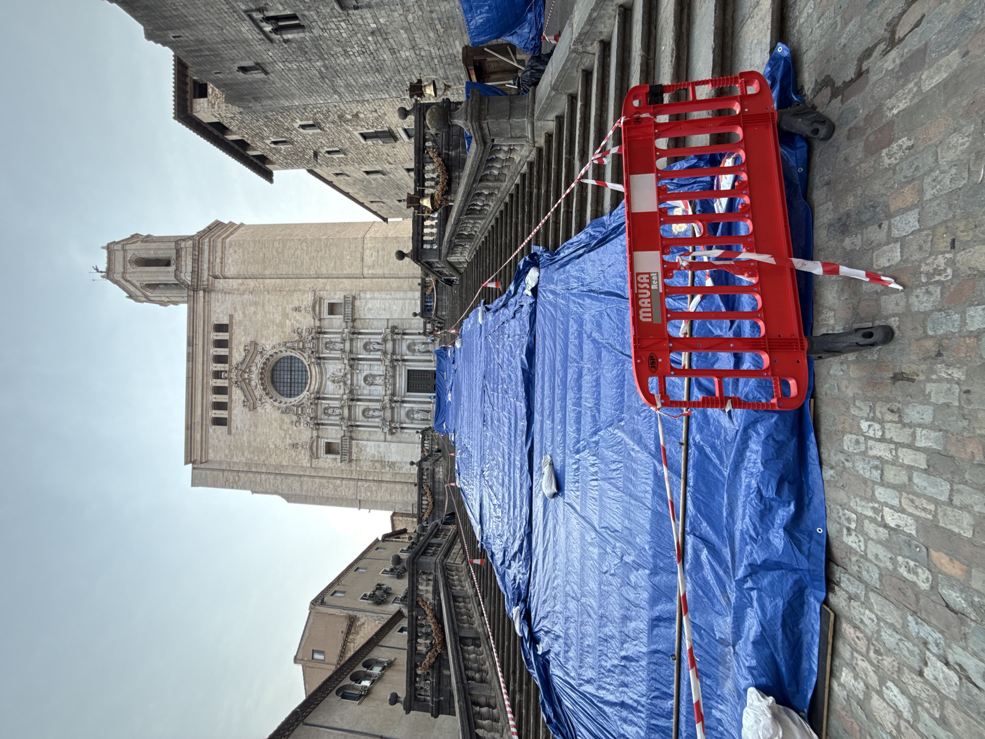 The image depicts a wide stone staircase leading up to a grand stone building, identified as the Girona Cathedral in Girona, Spain. The staircase and surrounding area are partially covered with large blue tarpaulins, suggesting ongoing construction or maintenance work. Red and white striped caution tape is strung across sections of the tarpaulin and stairs, and a red portable barrier with the logo "MAUSA RENT" is placed in the foreground, further indicating restricted access. The architecture of the cathedral is visible at the top of the stairs, featuring a large rose window and ornate carvings. Adjacent buildings, also constructed of stone, frame the scene. The sky is overcast. There are no visible people in the image.