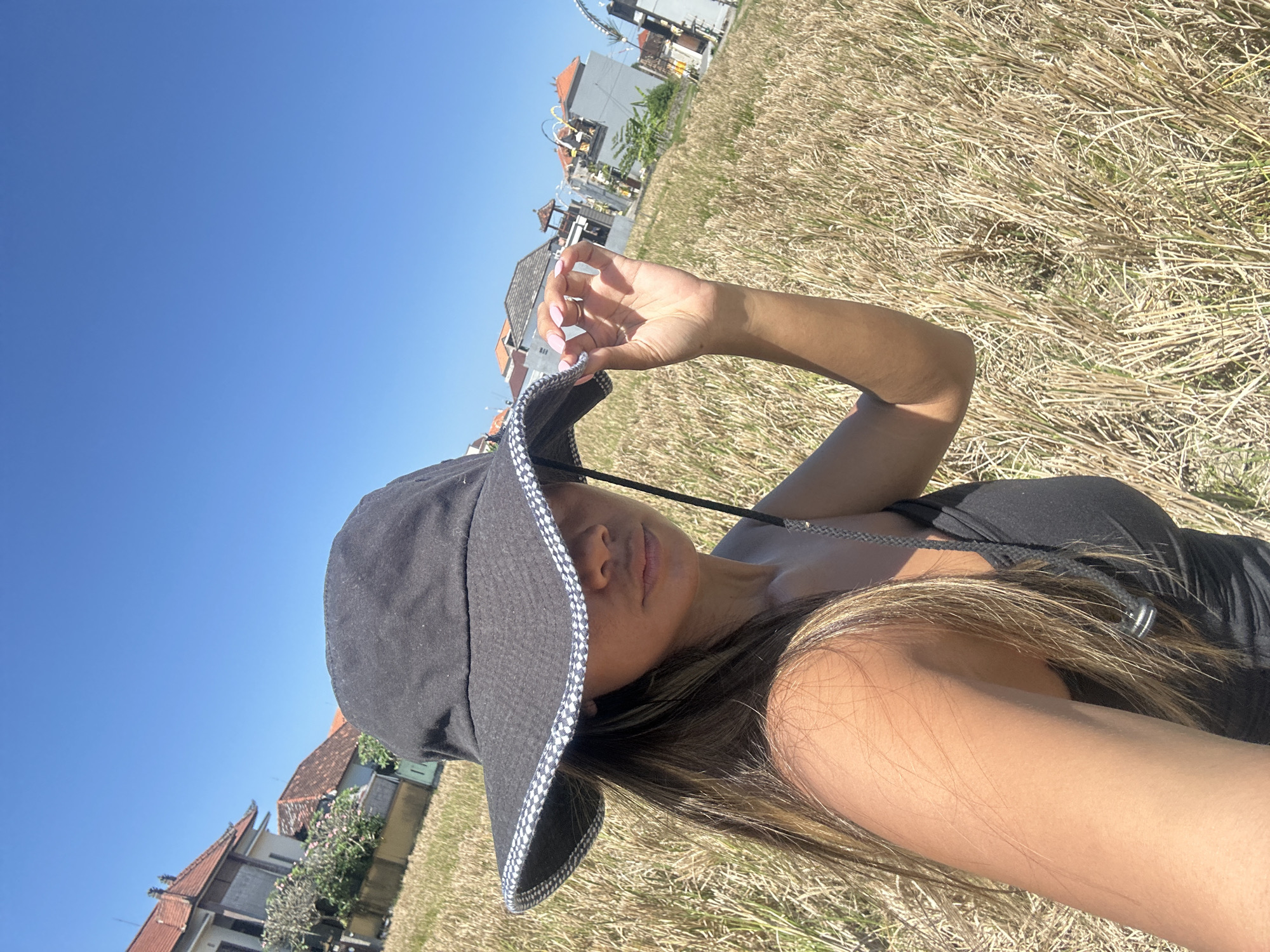 The media file shows a woman lying down in a dry, grassy field under a bright blue sky. She is wearing a black bucket hat with a checkered brim and a black tank top. Her right hand is raised, touching the brim of her hat, and her left arm is resting on her chest. Her long, brown hair is spread out on the grass. In the background, there are buildings with traditional Indonesian architecture, characterized by their tiled roofs and ornate details. The scene appears to be set during the daytime, as indicated by the clear sky and strong sunlight casting shadows. The dry grass suggests a warm, possibly dry season. The overall impression is one of relaxation and enjoying the outdoors in a rural or semi-rural setting. There is no visible text in the media file.