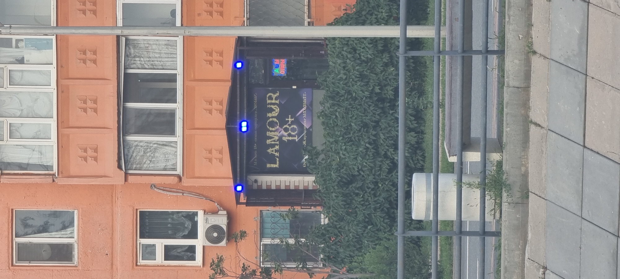 The image captures a storefront with a prominent sign reading "LAMOUR 18+". The storefront is located on the ground floor of an orange-colored building. Blue lights illuminate the entrance area, and a smaller sign with red lettering that says "ОТКРЫТЫ" (Open) is visible to the right. The building has multiple windows with curtains. Lush green bushes obscure the lower part of the storefront. In the foreground, a metal railing runs horizontally, separating the street from a concrete area with a bench and a cylindrical concrete object. The overall impression is of an urban commercial area in Aktobe, Kazakhstan, with a focus on the establishment's signage.