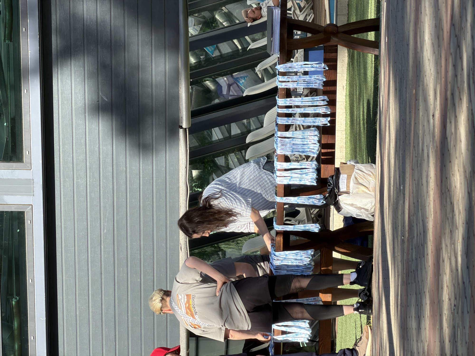 Two women stand at a wooden table outdoors. A large building is in the background, and sunlight casts strong shadows. The table is stacked with piles of blue and white ribbons. One woman wears a grey shirt, black leggings, and black shoes. The other wears a striped blue and white shirt.