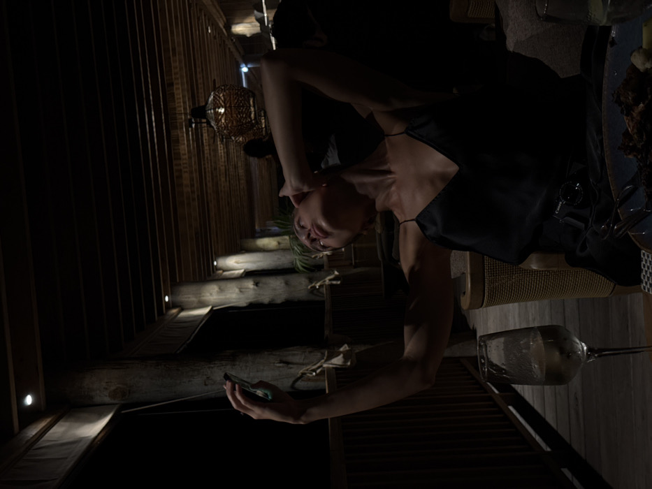 A woman in a black dress uses her phone on a wooden patio. String lights illuminate the wooden structure. A wine glass sits on a table next to a plate with food. The image is shot at a rotated angle.