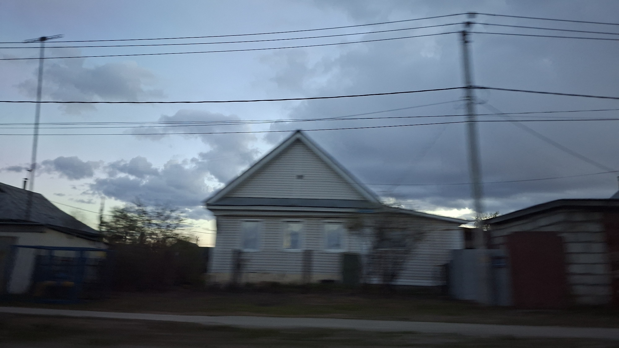 The image shows a one-story house with a gabled roof in a residential area. The house has light-colored siding and several windows. An electrical pole and power lines are visible against a cloudy sky. The image has a somewhat blurry, unclear quality. Based on the prompt, this is likely in Chaikovsky, Russia. The house appears to be a modest dwelling, perhaps a single-family home. The surrounding area seems to be a quiet neighborhood with simple houses. The lack of people suggests a potentially deserted time or place, but the presence of power lines indicates the availability of amenities.