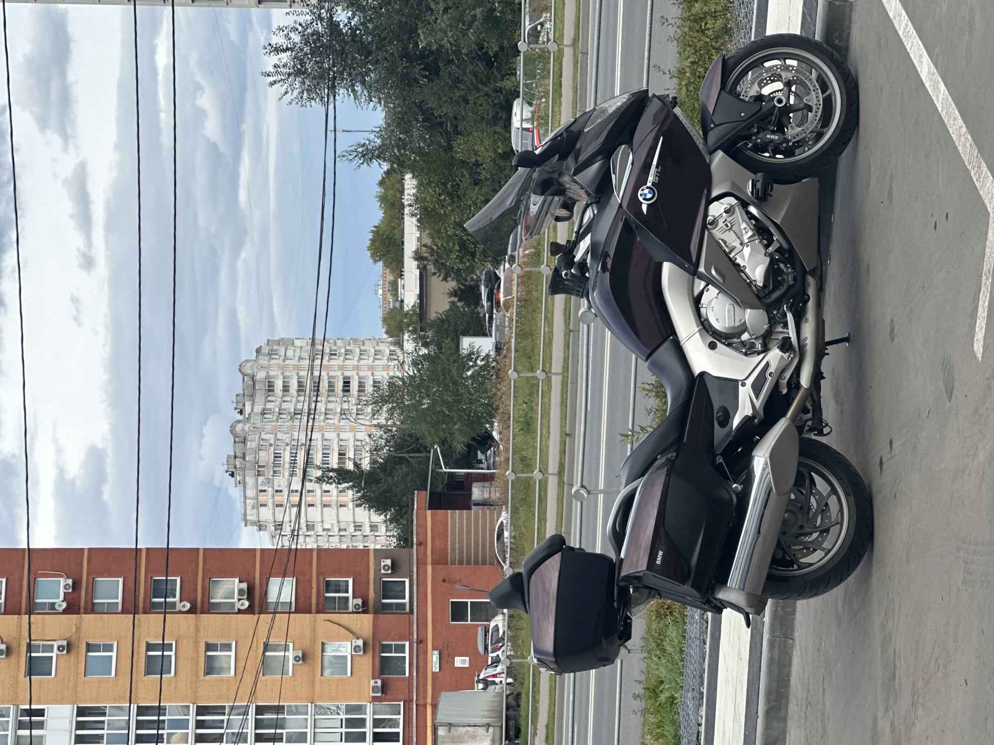 This is a medium-shot, eye-level, outdoor photograph of a dark red and silver BMW K1600 GTL motorcycle parked on a paved surface. The motorcycle is oriented diagonally, facing towards the right side of the frame, with its front wheel slightly turned. It features large saddlebags and a top case. The background depicts an urban scene in Podolsk, Russia. To the left, there is a tall, multi-story residential building with alternating orange and red brickwork and numerous windows and air conditioning units. Further in the background, a complex of similar apartment buildings is visible under a cloudy sky. A metal fence runs horizontally across the middle ground, separating the motorcycle's parking area from a road where several cars are visible. Power lines are strung across the upper portion of the image. The lighting suggests an overcast day.