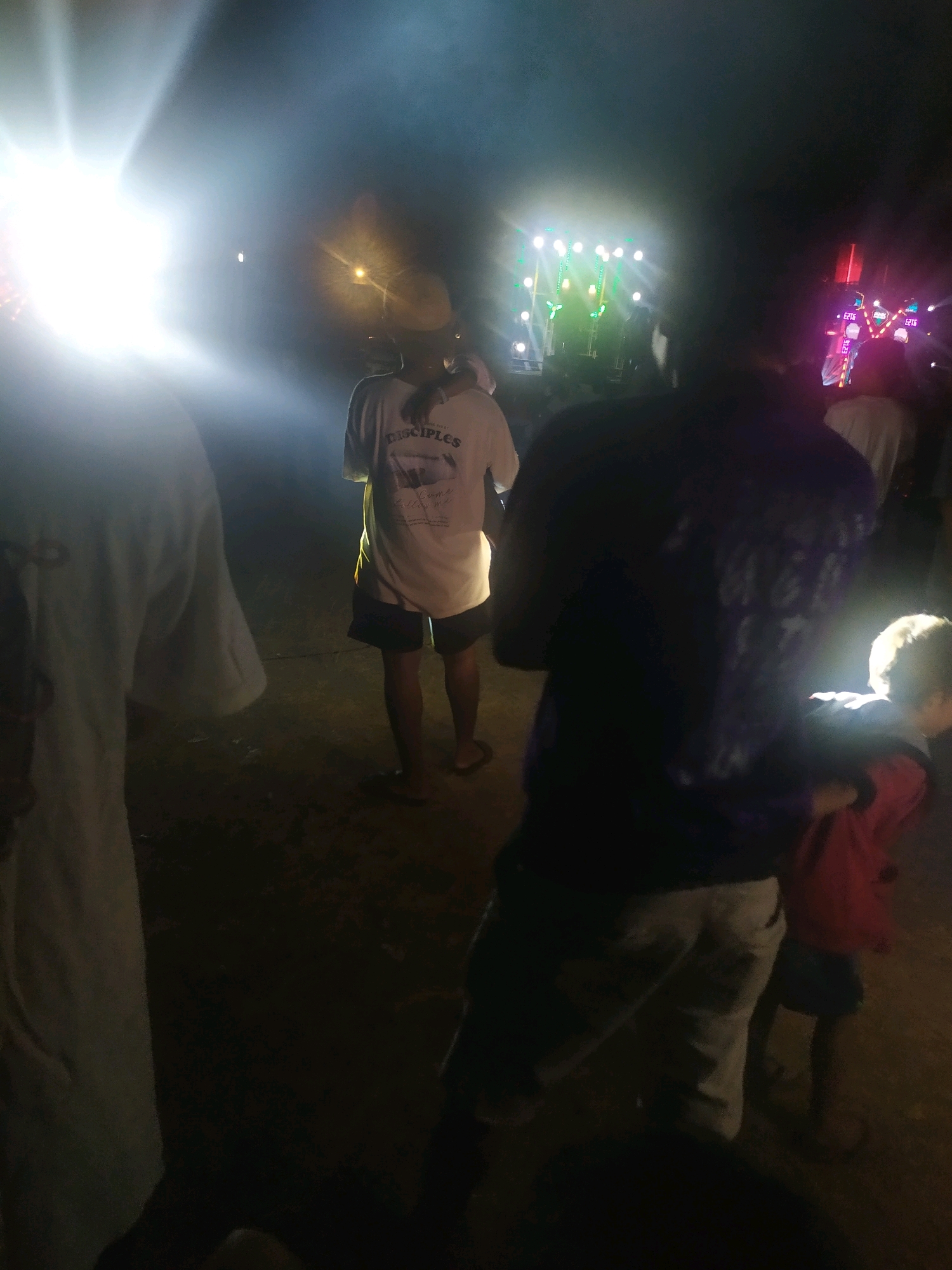 This image, taken at night in Gempol, Indonesia, depicts a gathering illuminated by bright stage lights. In the foreground, a person wearing a white t-shirt stands with their back to the camera, their t-shirt bearing the word "DISCIPLES." To their left, another individual in a white t-shirt is partially visible. To the right, a person in a dark purple shirt and light-colored pants stands, with a child in a red jacket next to them. In the background, a stage with multiple lights and what appears to be sound equipment is illuminated. The ground is visible and appears to be dirt or gravel. The atmosphere suggests an outdoor event, possibly a concert or festival, with a significant crowd present.