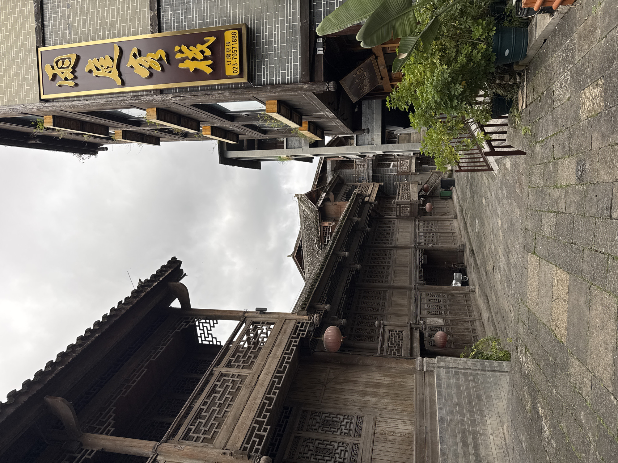 The image depicts a street scene in Zhuoshui, China, characterized by traditional wooden architecture with intricate latticework and overhanging eaves. The buildings are constructed from dark wood and gray brick. A sign with gold Chinese characters is visible on one building, indicating a business or establishment. Red lanterns hang from the eaves of one of the buildings. The street is paved with gray stone slabs. The sky is overcast.