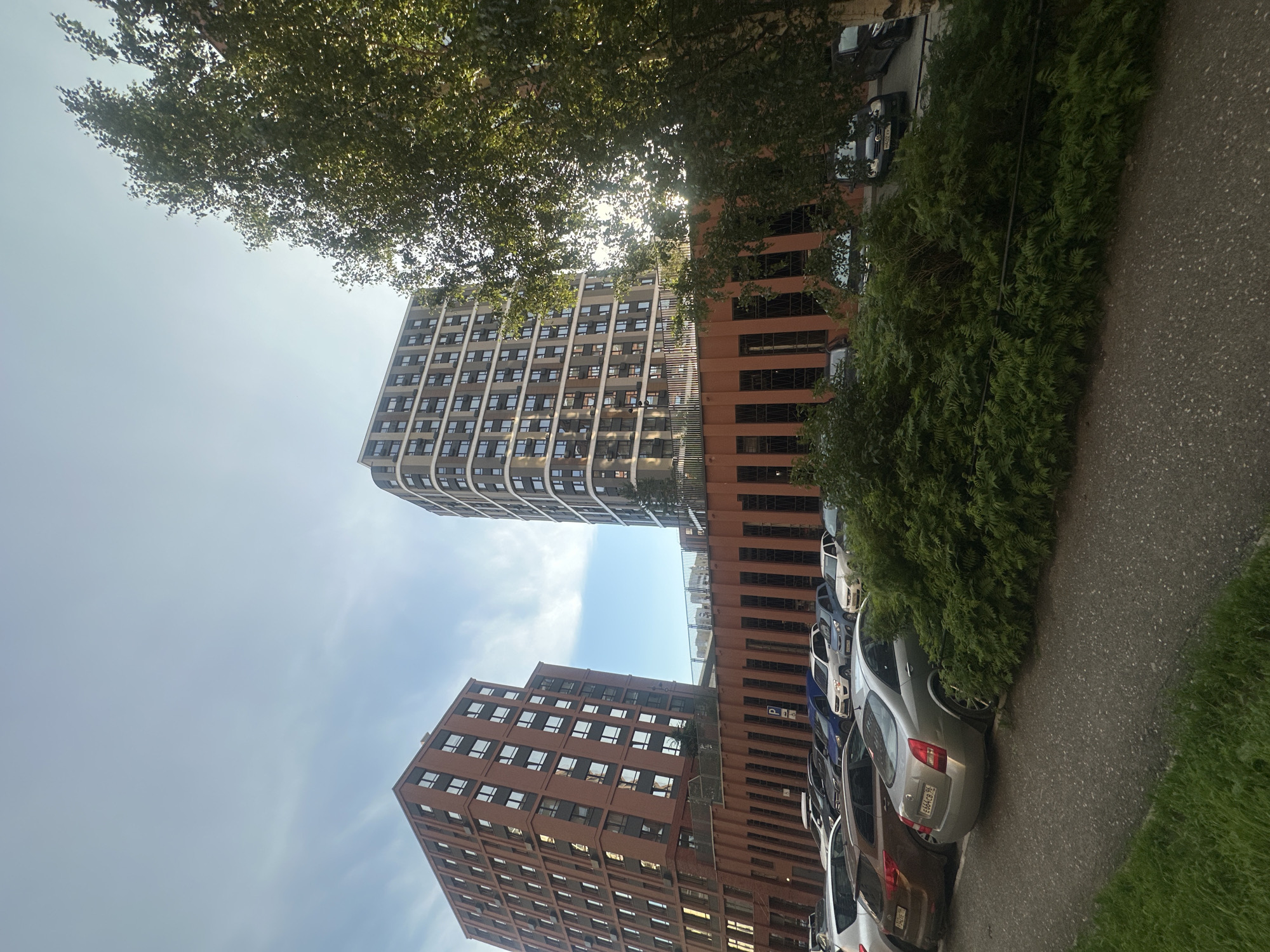 This image, captured in Yekaterinburg, Russia, shows a street view with modern apartment buildings and parked cars. The buildings are contemporary in design, with one being a tall, rectangular structure clad in red brick and featuring numerous windows. Next to it stands another taller building with a curved facade, also adorned with many windows. In the foreground, a row of parked cars, including sedans and SUVs, are visible. A dense cluster of green foliage obscures some of the vehicles and a black fence. The sky above is blue with some wispy clouds, and the sun appears to be shining, casting light and shadows. The scene depicts a typical urban residential area.