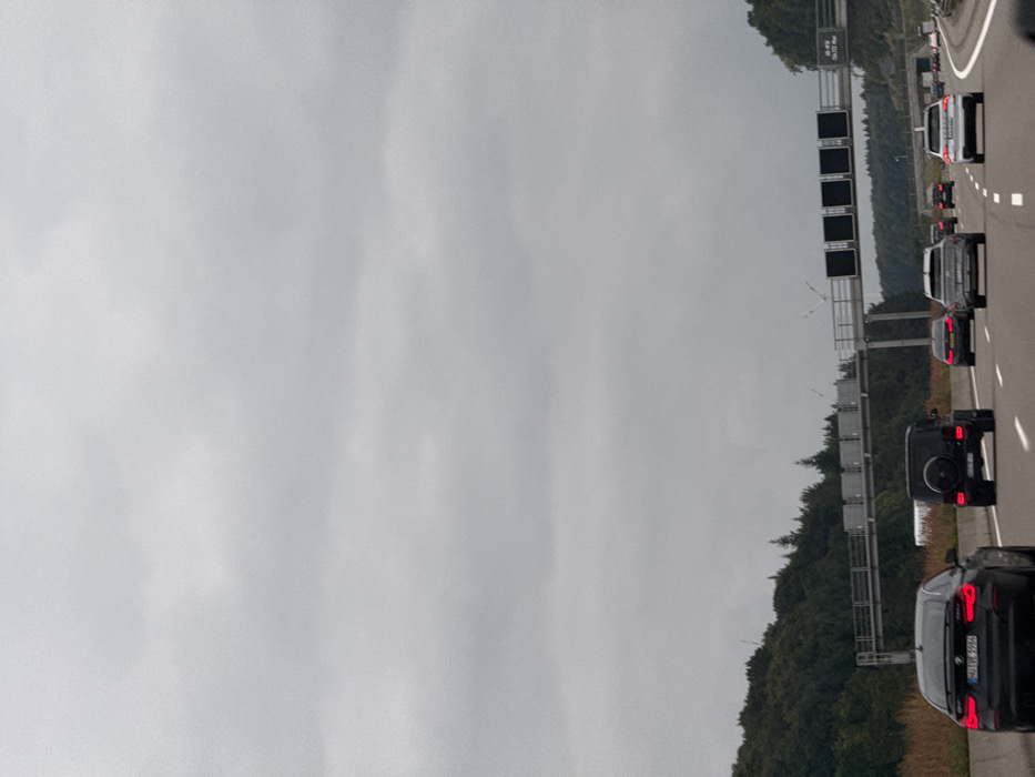 The image displays a multi-lane segment of the German Autobahn 8 (A8) near Dornstadt, Germany, under a uniformly overcast sky. The roadway features multiple lanes of traffic, with vehicles proceeding in the same direction as the viewer. In the immediate foreground, a black BMW 4 Series Gran Coupé, bearing the German license plate `HD HV 9556`, occupies the left lane. Ahead of it, a black Mercedes-Benz G-Class SUV is positioned in the middle lane. Further down the road, several additional vehicles are observed, including a gray Audi SUV and a white SUV in the right lane, and a dark sedan in the left lane. An overhead gantry spans the highway, supporting multiple black, unlit variable message signs and a fixed sign on the right side displaying "A8 - PÖL..." and a kilometer marker "136,777 km". Dense forest, composed of both deciduous and coniferous trees, lines the elevated terrain flanking both sides of the highway. The sky is entirely covered by a thick layer of gray clouds. No individuals are visible.