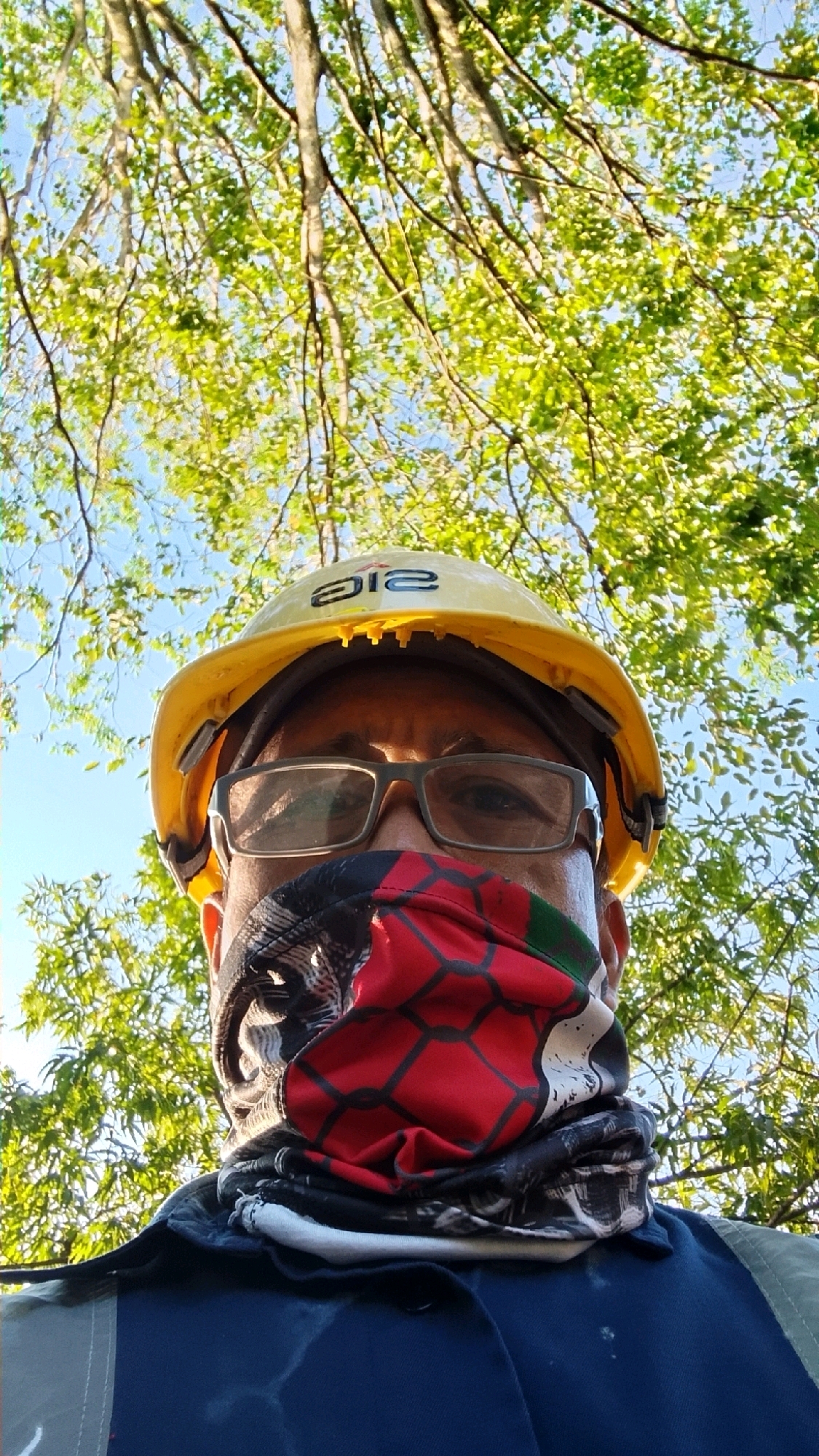 A low-angle, eye-level, medium shot captures a person wearing a yellow hard hat, glasses, and a multi-colored face covering. The person is wearing a dark blue shirt and is partially obscured by trees with green leaves and a bright blue sky in the background. The hard hat has a black logo with white lettering. The face covering is red with black geometric patterns and some green at the top. The individual's eyes are visible through the glasses. The overall scene appears to be outdoors, possibly in a park or wooded area. The provided metadata suggests the location is Tlogobendung, Indonesia.