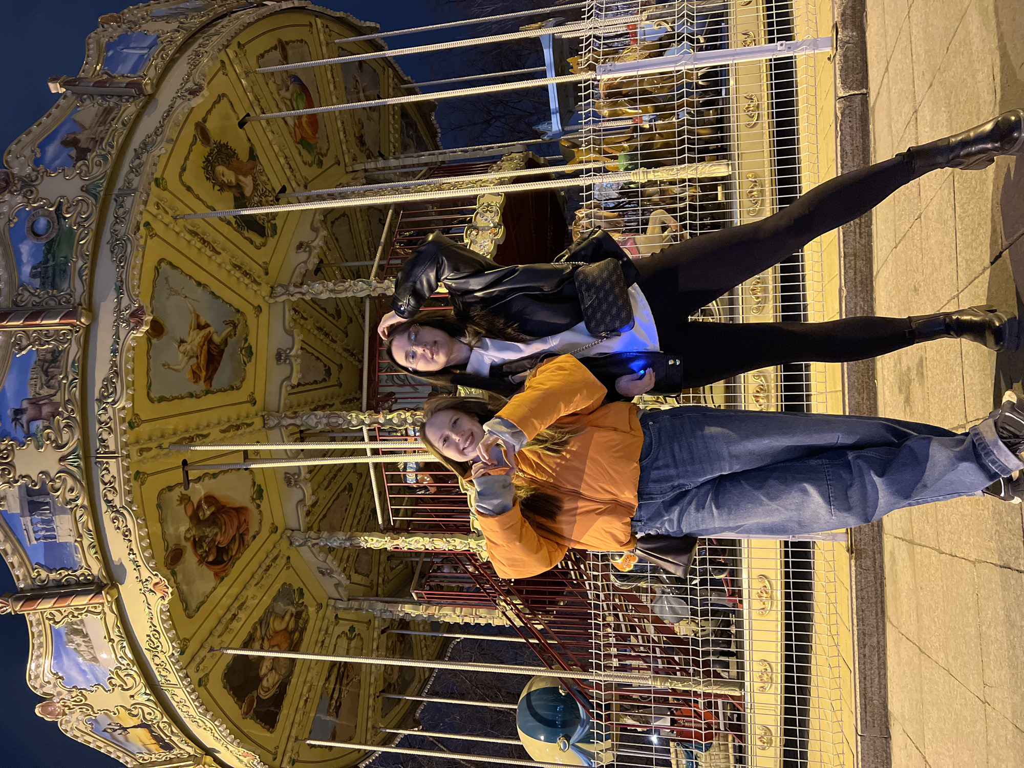 Two young women pose in front of a carousel in Tyumen, Russia. One wears an orange jacket and jeans, making a heart shape with her hands. The other wears a black leather jacket and leggings. The carousel has a highly decorated canopy with paintings and ornamental details. A safety barrier surrounds the carousel. It appears to be evening, with artificial lighting illuminating the scene.