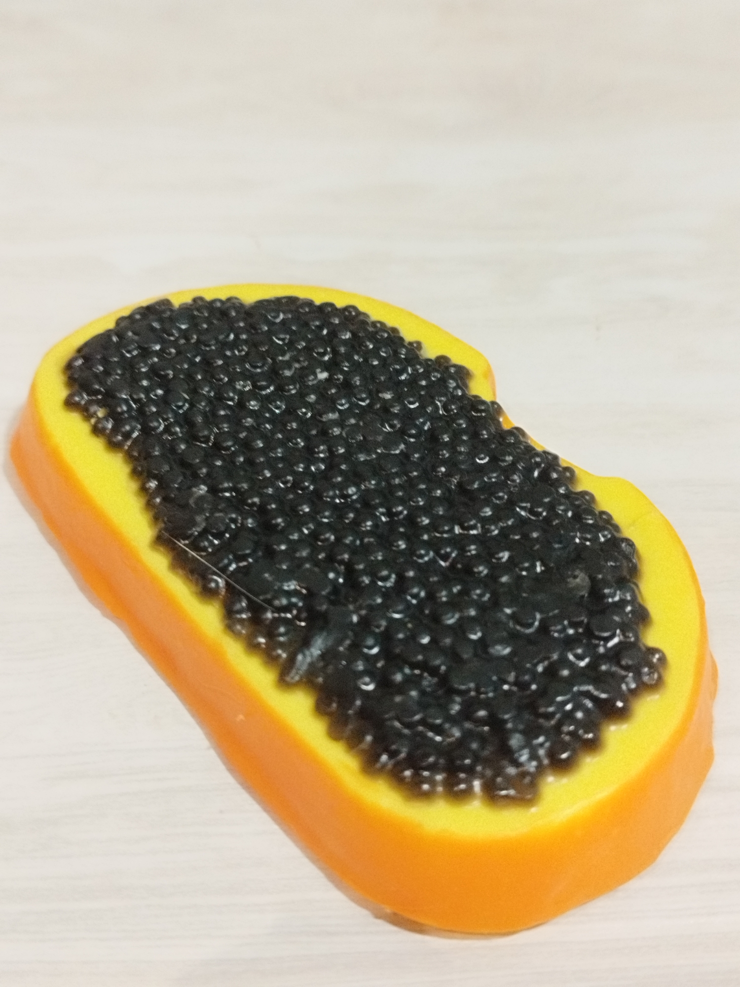 A close-up, overhead shot shows a plastic toy resembling a slice of papaya topped with black caviar, resting on a light-colored, wood-grain surface. The papaya slice is orange on the rind and yellow on the fruit, with a textured layer of small, black, caviar-like beads covering the yellow section. The toy is positioned diagonally, with the rind facing the bottom right and the caviar-laden portion extending towards the top left. The lighting is even, casting no strong shadows. The scene is devoid of any human presence or any indication of a specific location within Saint Petersburg, Russia.