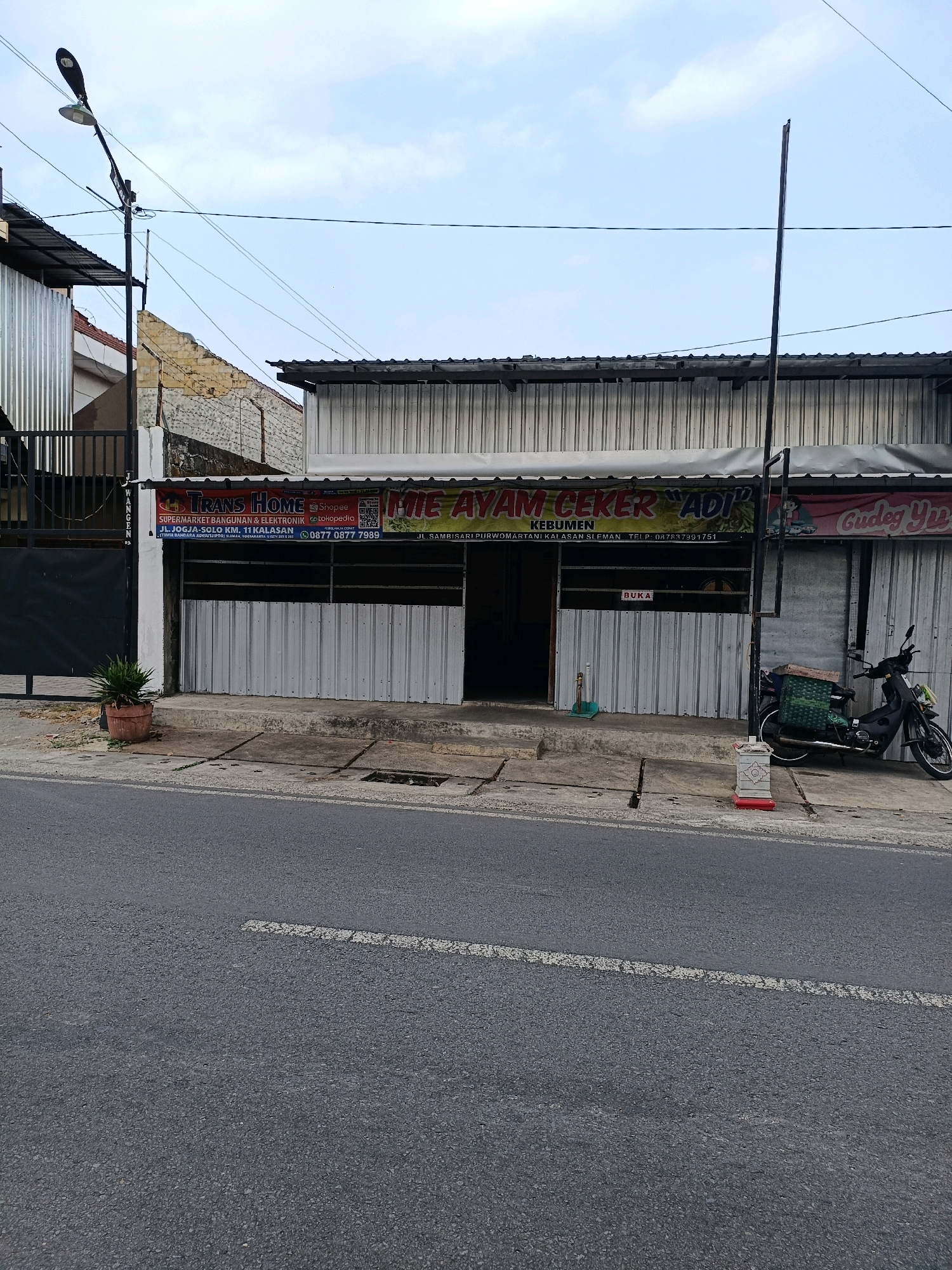 The image shows a street view in Purwomartani, Indonesia, with a row of buildings lining the right side of the road. The buildings have metal siding and awnings. A sign for "TRANS HOME SUPERMARKET BANGUNAN & ELEKTRONIK" is visible on the left building. To the right, a sign for "THE AYAM CEKER ADI" and "Gudeg Yuy" are also displayed, indicating food establishments. A black motorcycle is parked in front of one of the buildings. A street lamp is positioned on the left side of the image, and power lines are visible overhead. The road in the foreground is paved with asphalt and has a white line marking. A potted plant sits on the sidewalk near the left building. The sky is partly cloudy.