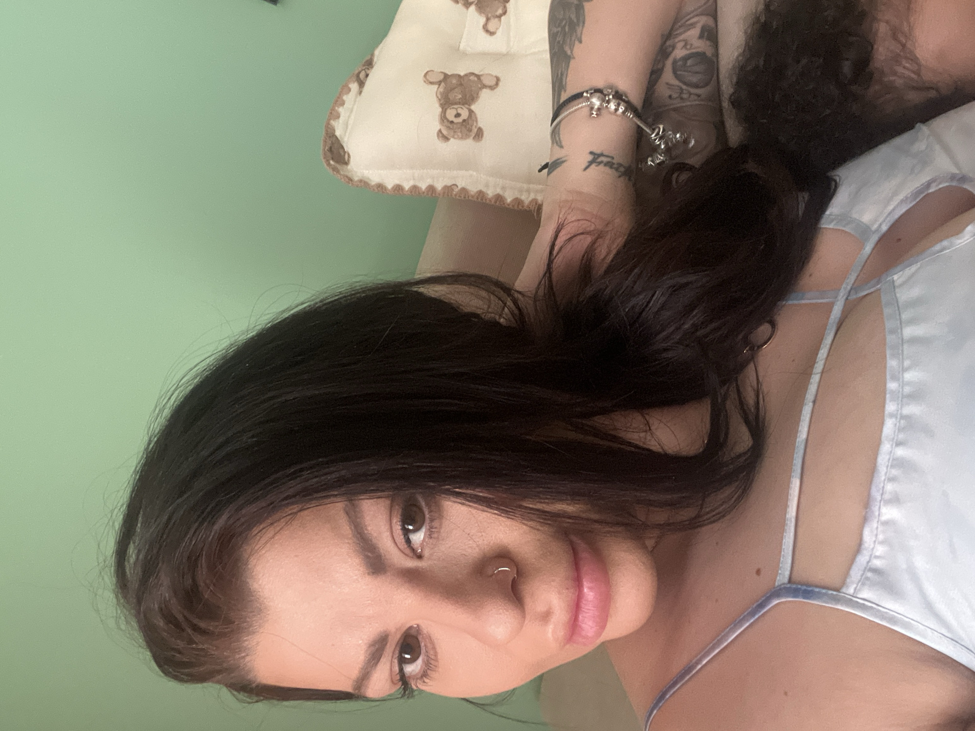 The image is a close-up selfie of a woman with dark hair and light-colored clothing, taken indoors. The woman is lying down, with her head slightly tilted. She is wearing a light blue, spaghetti-strap top. A pillow with a teddy bear pattern is visible behind her. Her left arm is resting on the pillow, and she is wearing a bracelet and several tattoos are visible on her arm. The background is a solid green wall. There is no indication of the location being in Plovdiv, Bulgaria.