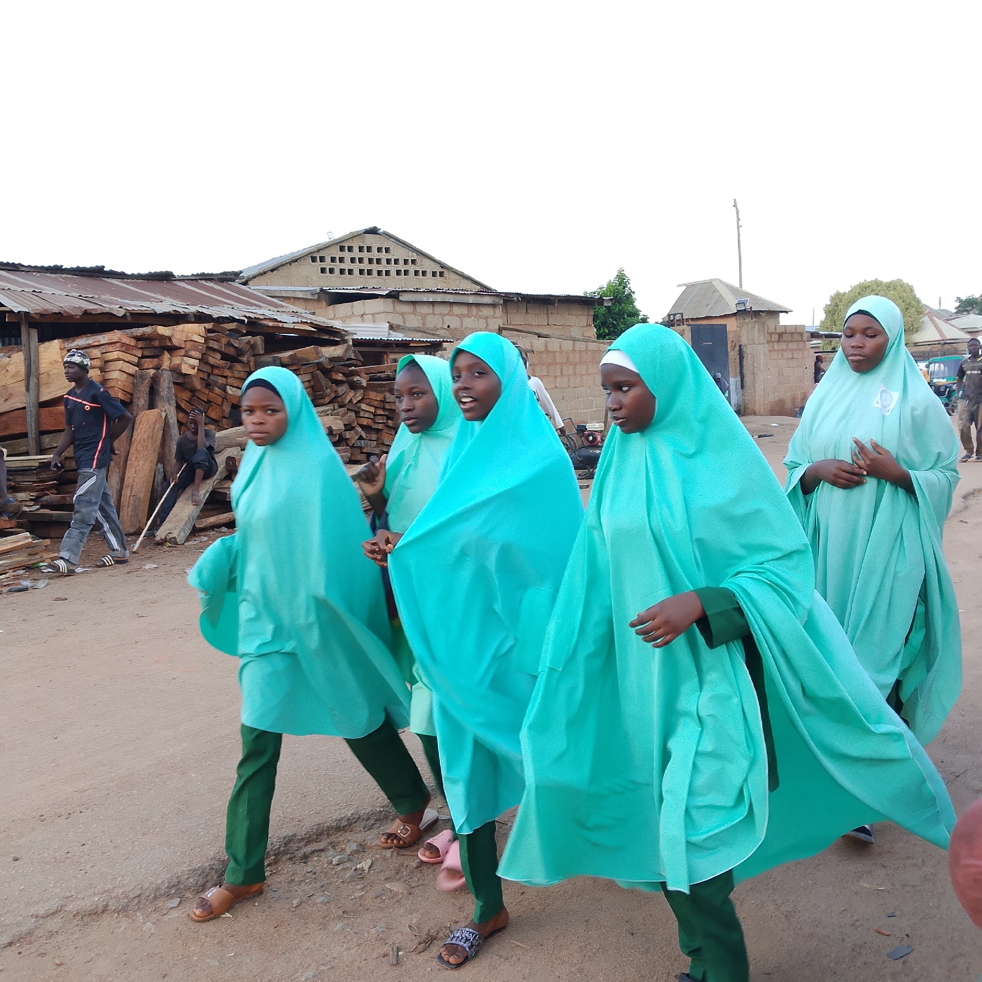 In the city of Unknown, Unknown, five young girls, dressed in teal hijabs and green trousers, are walking on a dirt road. They are walking from left to right, with the girl in the foreground on the right facing the camera. Behind them, to the left, a young man in a black shirt and grey trousers walks away from the camera. Further in the background, several buildings with corrugated iron roofs and brick walls are visible, along with a large stack of lumber. The sky is overcast.