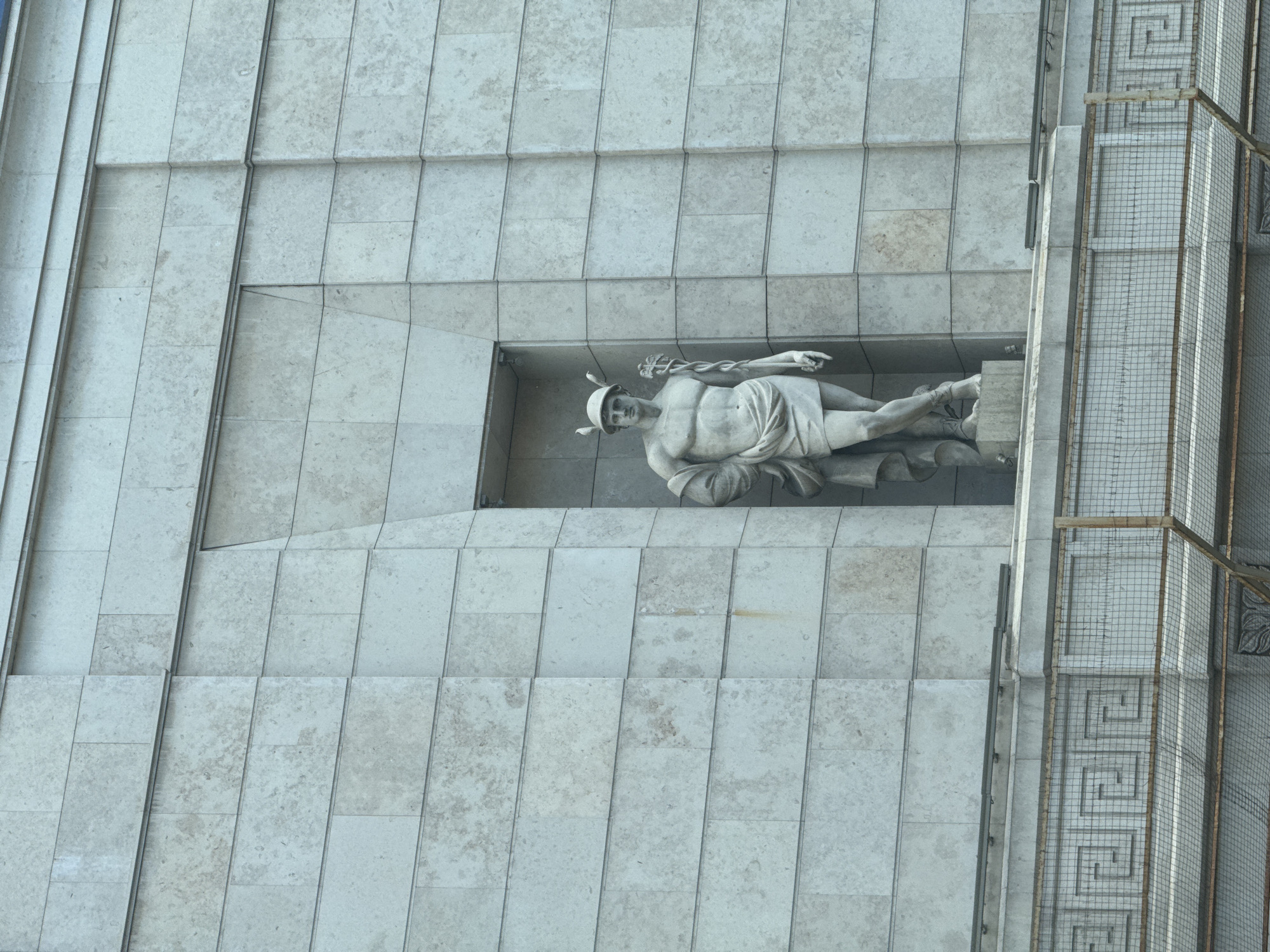 This is a vertically oriented, close-up shot of a monumental sculpture of a winged male figure, possibly Mercury or an angel, set within a recessed niche on the exterior of a modern, grey stone building. The sculpture is rendered in a classical style, with the figure reclining on its left side, looking upwards and to the left. The figure is draped in a loincloth and wears a winged helmet and sandals. A caduceus, a symbol often associated with Hermes/Mercury, is held in its right hand. The building's facade is composed of rectangular stone tiles arranged in a grid pattern. To the right of the niche, a section of the building features a decorative element with a Greek key motif and is covered by a wire mesh fence, suggesting ongoing construction or maintenance. The lighting suggests it is daytime, with no strong shadows, indicating an overcast sky or diffuse light. The overall impression is one of grandeur and stillness.