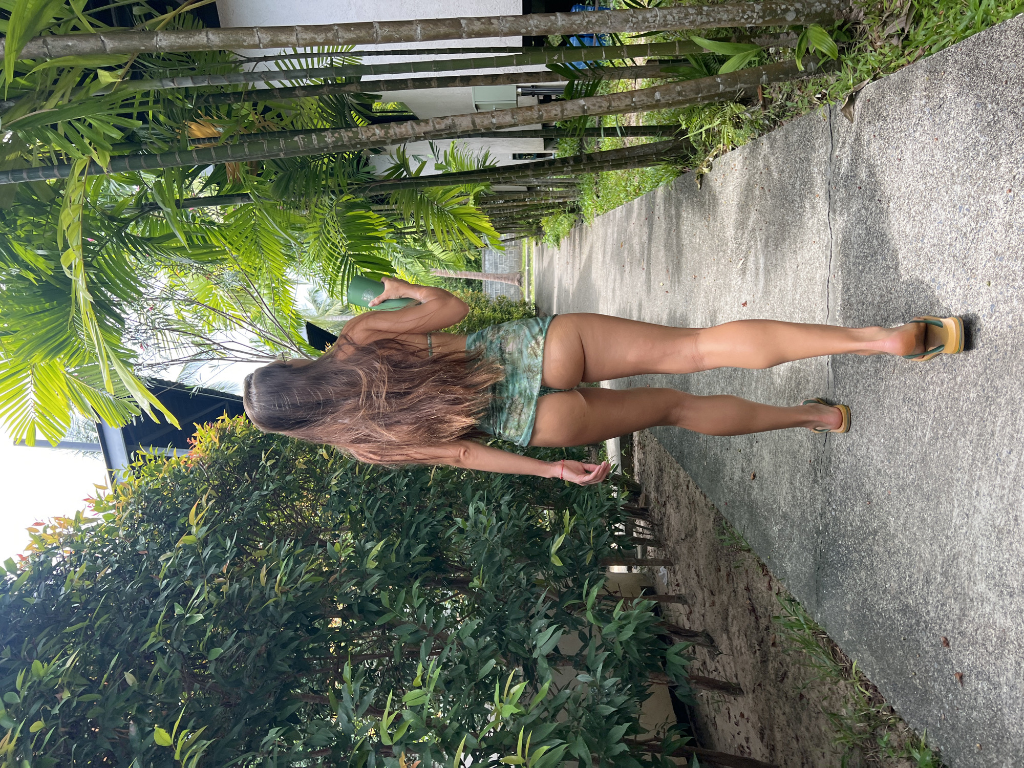A woman with long brown hair is walking away from the camera down a concrete path lined with lush green foliage and tall bamboo stalks. She is wearing a green patterned bikini and flip-flops. In her right hand, she holds a green cup. The scene appears to be a tropical resort or garden path in Baan Bang Rak, Thailand, characterized by the dense tropical vegetation and the architectural style of the buildings visible in the background.
