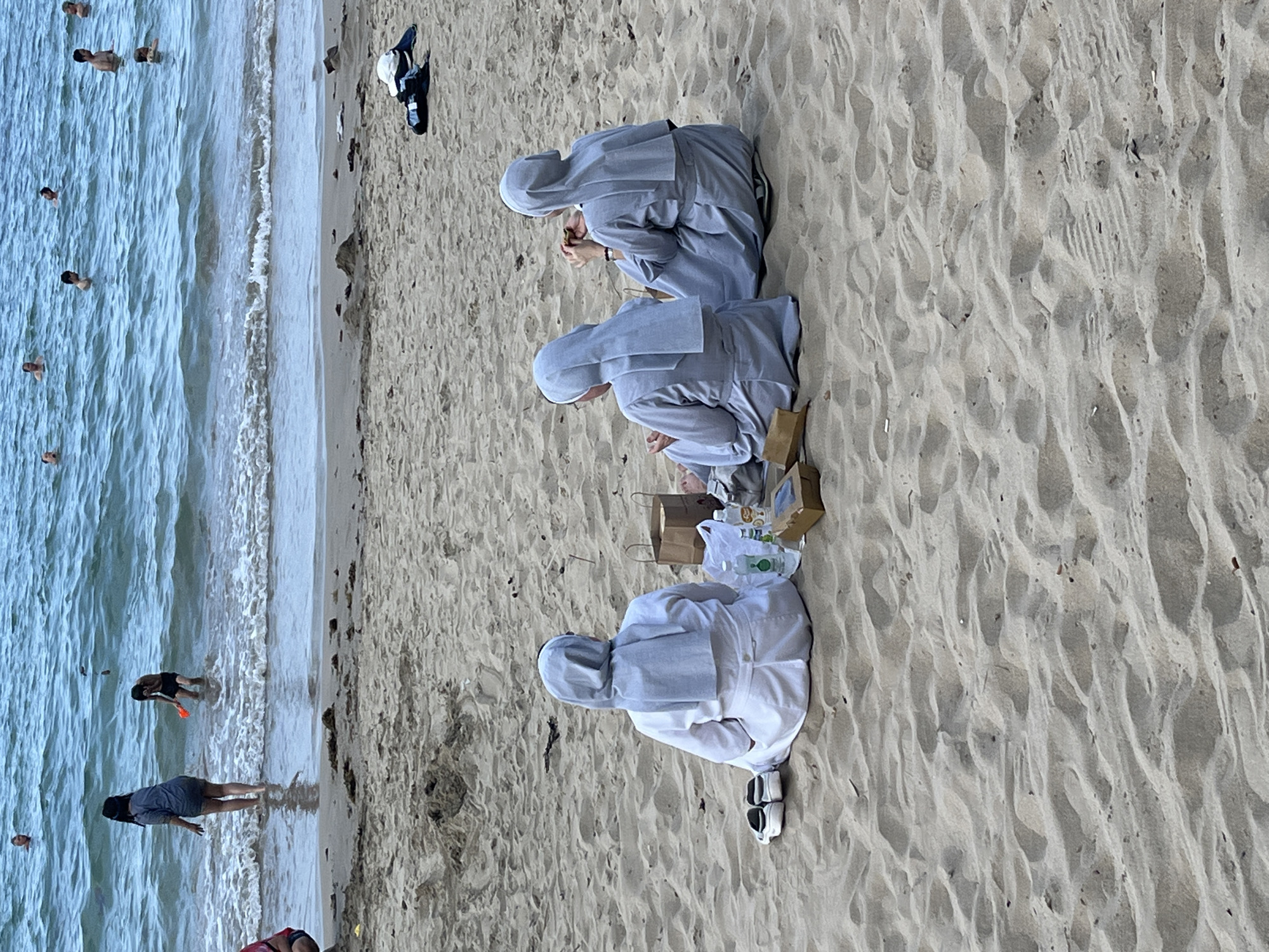 Three nuns in gray and white robes are seated on the sandy shore of a beach, possibly in or near Bắc Nha Trang, Vietnam, based on the geography. They are situated close to each other, near some brown paper bags, food containers, and a water bottle, which suggests they are having a meal. The beach has a light-colored sand. The sea is visible to the left, with some people swimming or standing in the water. The weather appears to be clear. A few articles of clothing are abandoned on the sand.
