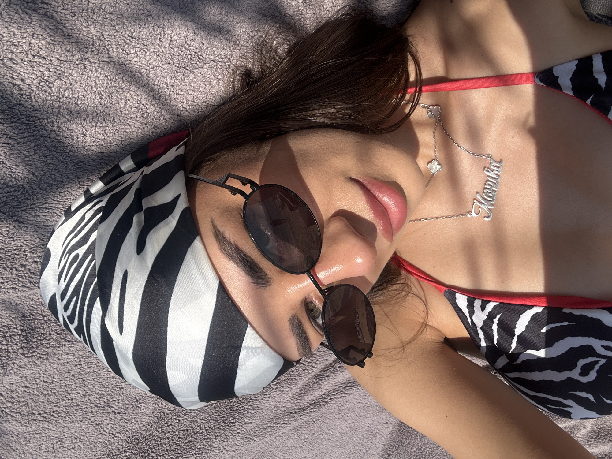 A young woman is lying on a towel outdoors, possibly at Ofrynio Beach, Greece, as indicated by the context. She is wearing a zebra-print bikini with red trim and a black and white zebra-print headscarf. She has on oval-shaped sunglasses and a silver necklace with a charm and her name, "Marilena", written in cursive. The woman is looking towards the camera with a slight smile. The sun is casting shadows on her face and the towel, suggesting it is daytime.