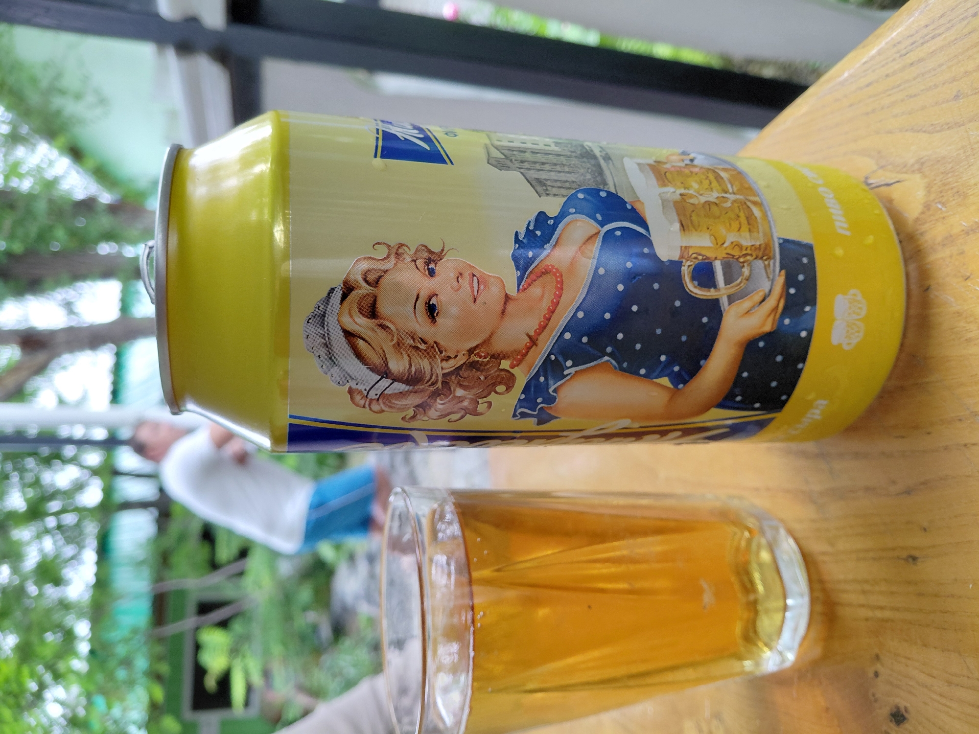 A close-up shot focuses on a yellow beer can with a vintage-style illustration of a woman in a blue polka-dot dress, holding a tray with two beer mugs. A glass of amber liquid, presumably beer, sits to the left of the can. The background is blurred, showing a person in a white t-shirt and blue shorts, suggesting an outdoor setting. The overall scene appears to be a casual, possibly outdoor gathering in a city location.