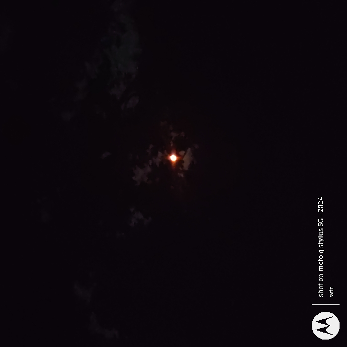 The image shows a full moon shining brightly in the night sky. The moon appears to be partially obscured by clouds. The image was taken in Happy Valley, Oregon, United States, at the coordinates 45.443, -122.552. The image was captured with a Motorola Stylus 5G phone in 2024. No people or other objects are visible in the image.